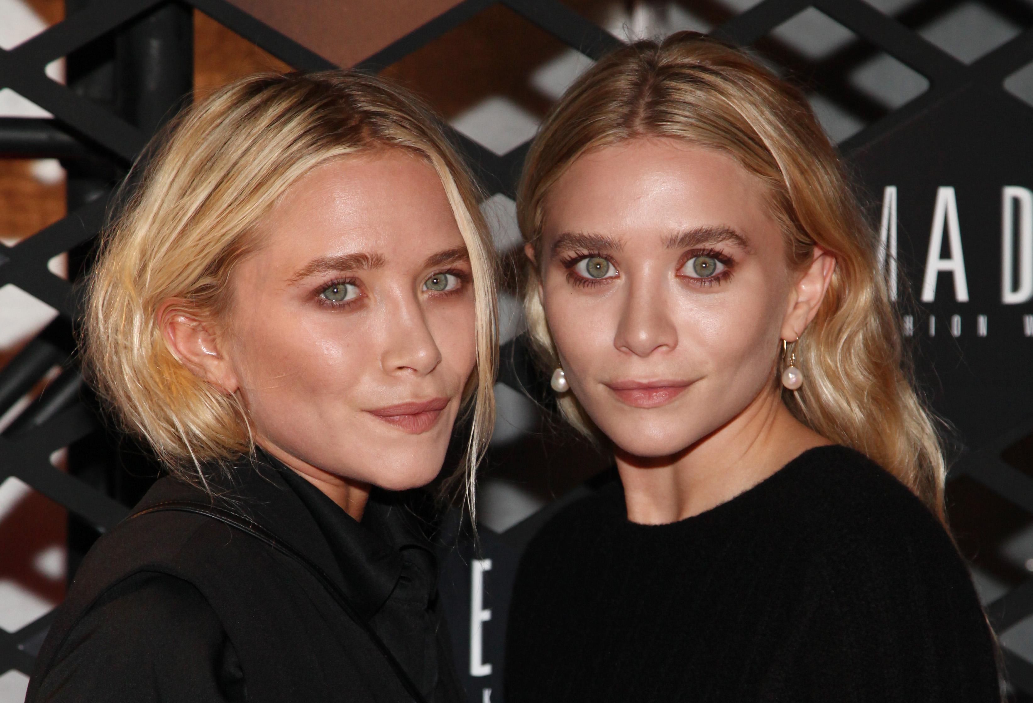 Mary-Kate and Ashley Olsen pose closely at a fashion event, facing the camera with calm expressions. Their minimal makeup and simple black outfits highlight their signature understated style.