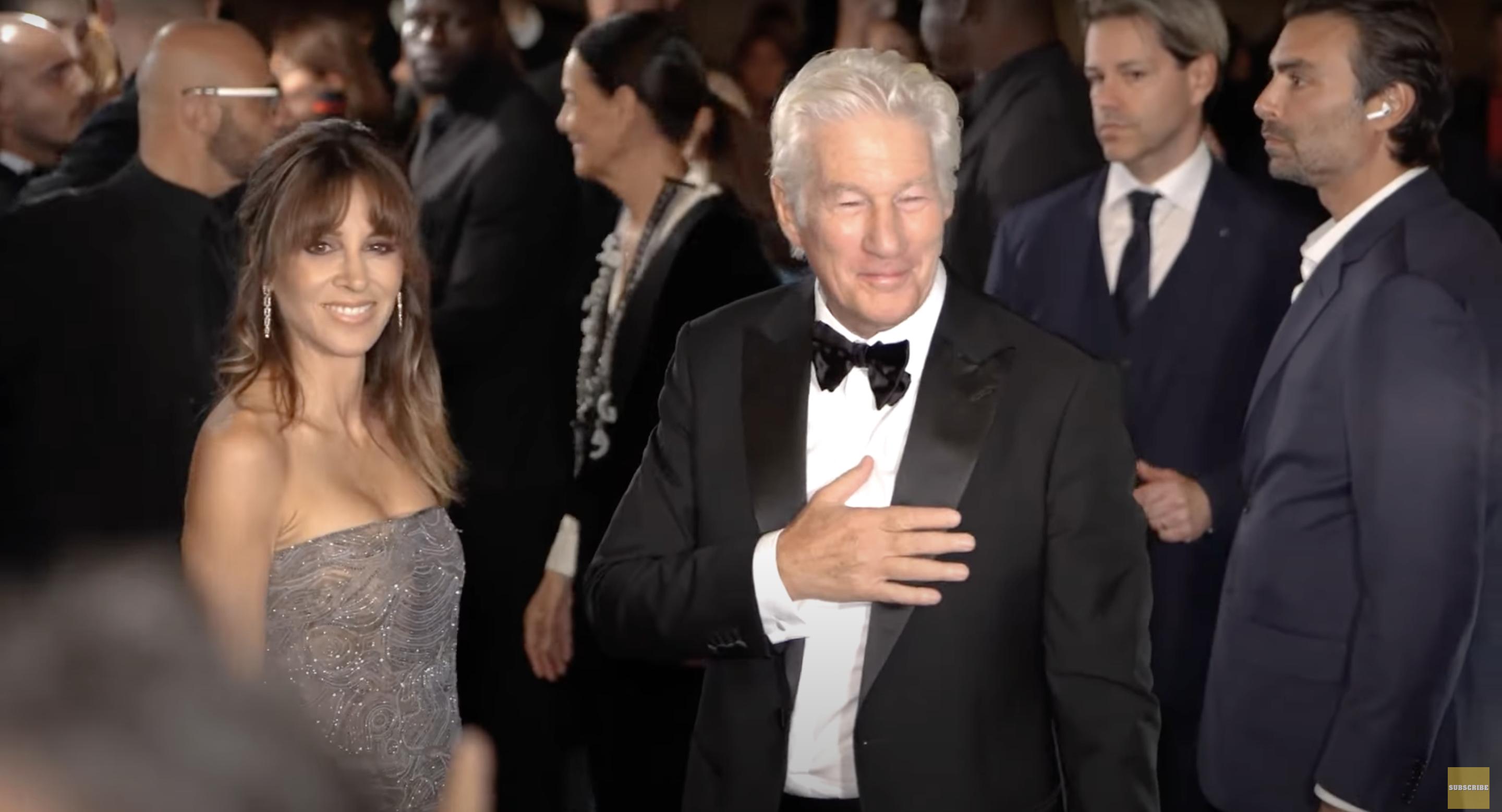 Alejandra Silva smiles confidently in a beaded silver gown, her hair styled in soft curls. Richard Gere stands beside her, hand over heart, in a classic black tux with a bow tie, appearing warm and composed. | Source: YouTube/pagesix