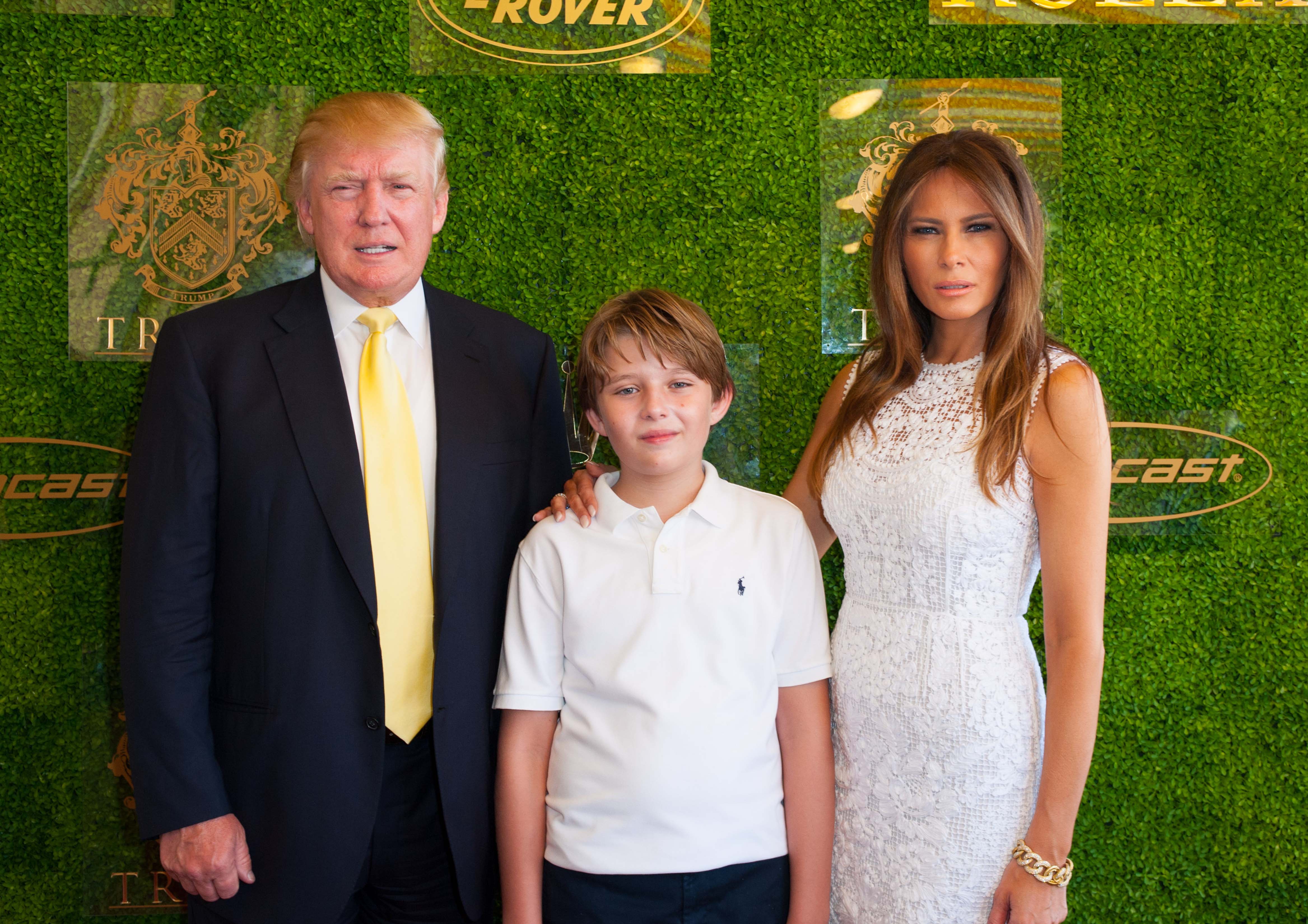 At the Trump Invitational Grand Prix in Palm Beach, Donald Trump stands with Melania Trump and their son, Barron Trump, before a green hedge backdrop. Melania wears a fitted white lace dress, while Barron appears in a white polo shirt and dark trousers. The trio pose closely together for photos at the Mar-a-Lago event.