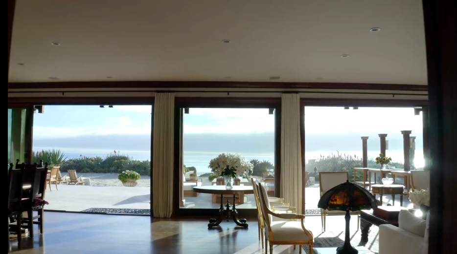Floor-to-ceiling glass sliding doors span the interior living space, creating a seamless connection to the outdoor terrace and ocean views beyond. Inside, refined furnishings like ornate gold-accented chairs, a classic table with a central floral arrangement, and a decorative stained-glass lamp elevate the room's warm aesthetic. | Source: YouTube/Architectural Digest
