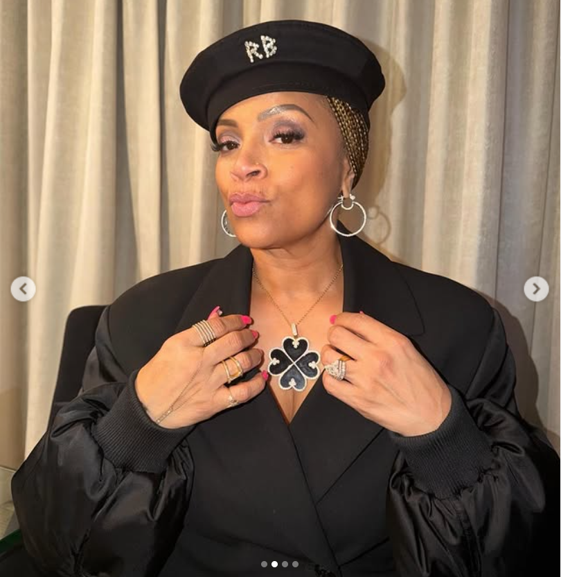 Simone Smith strikes a confident close-up pose, wearing a black beret with subtle embellishment, a tailored blazer, and bold silver hoop earrings, as she lightly adjusts a statement clover-shaped pendant necklace. | Source: Instagram/sislovespurple
