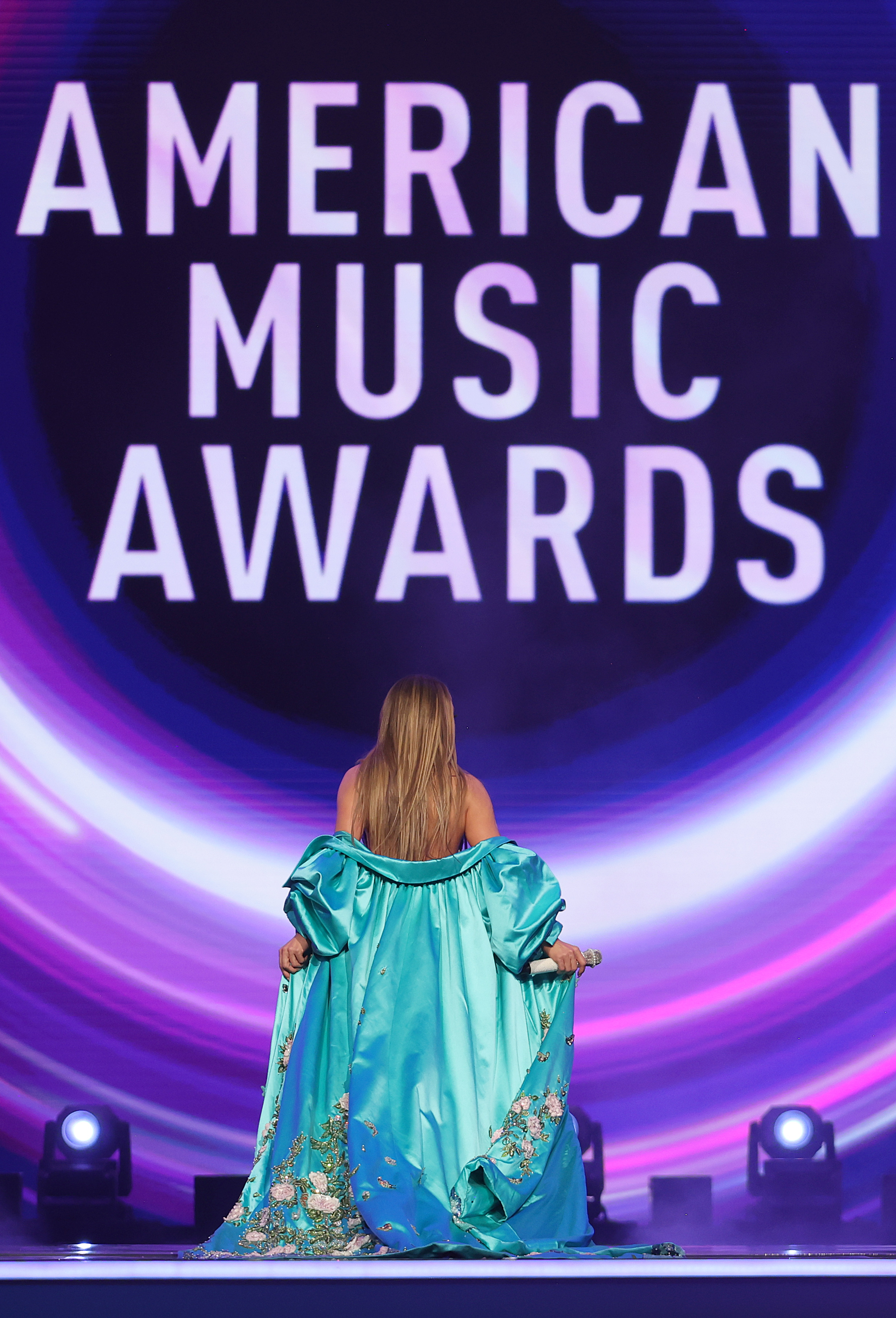 With the AMAs logo illuminated in front of her, Jennifer Lopez holds up her overcoat while standing center stage with a microphone in her hand.