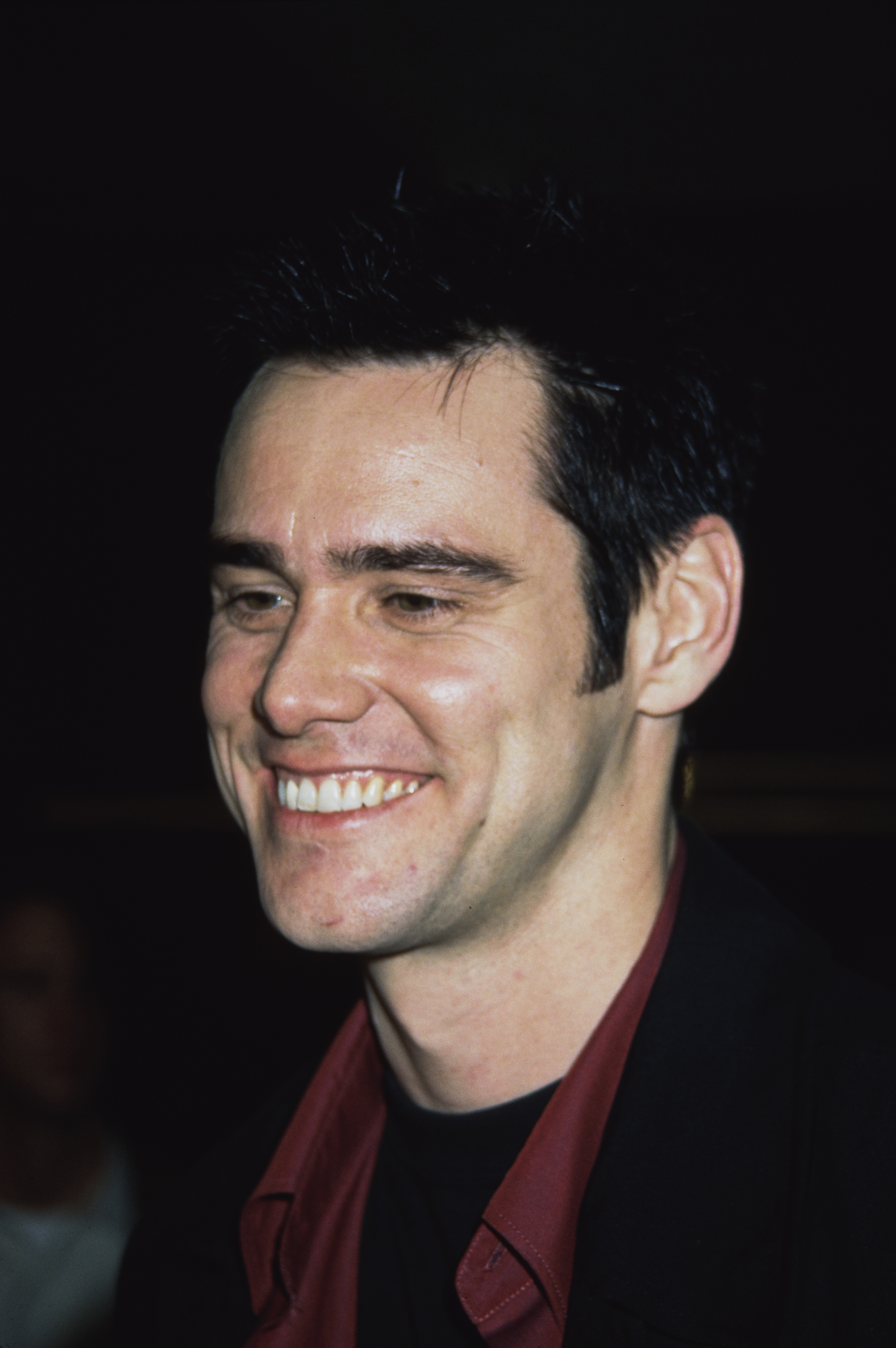 Jim Carrey is smiling warmly while attending the "Beautiful Girls" premiere at Cineplex Odeon Century Plaza Cinemas in California. Dressed in a black jacket over a maroon shirt, he exudes relaxed charm and confidence. The candid shot captures his friendly demeanor and natural charisma in front of the cameras.