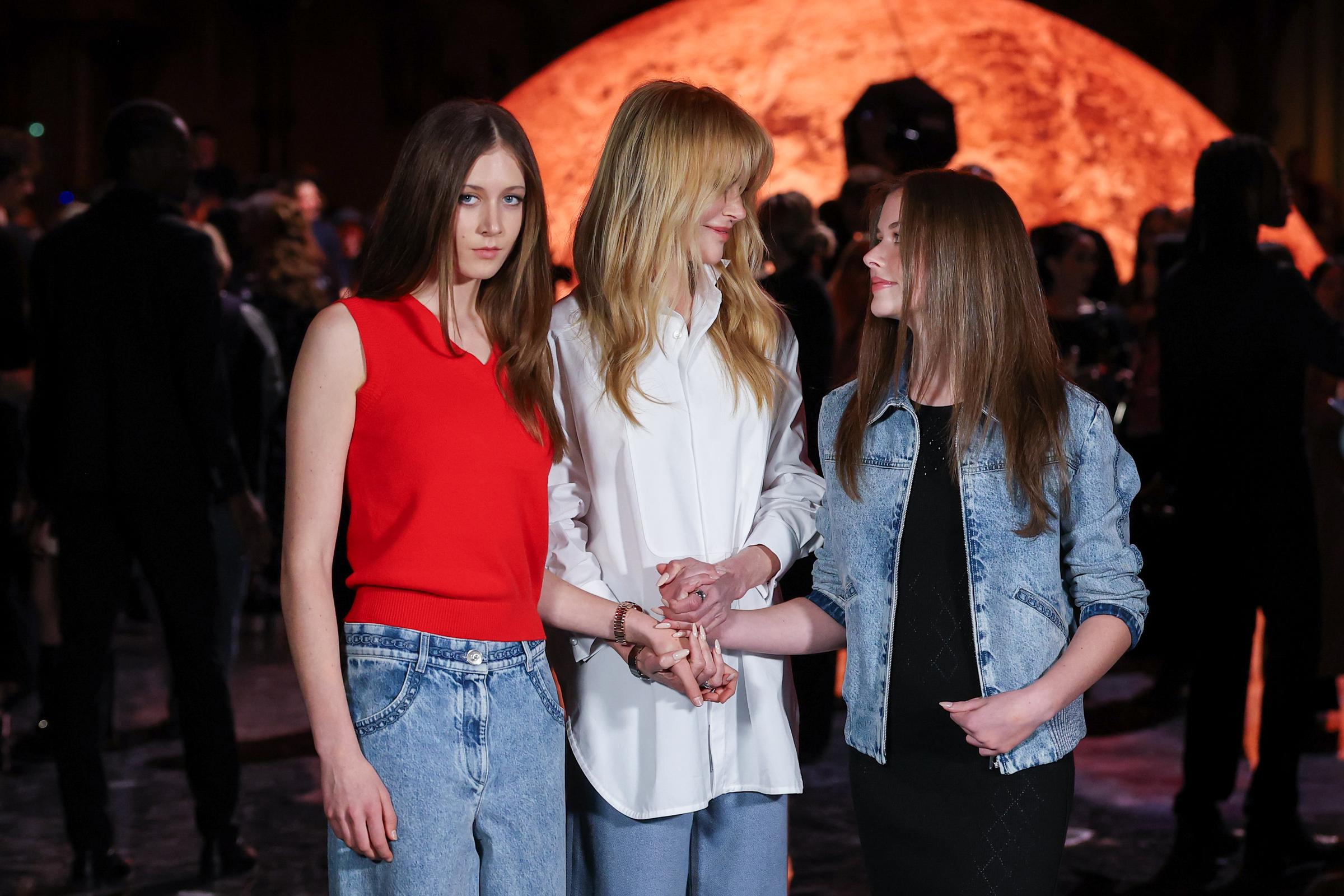 Nicole Kidman shares a warm moment with her daughters, Sunday Rose, and Faith Margaret, while attending the Chanel Womenswear Spring/Summer 2026 show during Paris Fashion Week, on October 6, 2025. The trio stands hand in hand under the soft glow of the event's dramatic lighting.