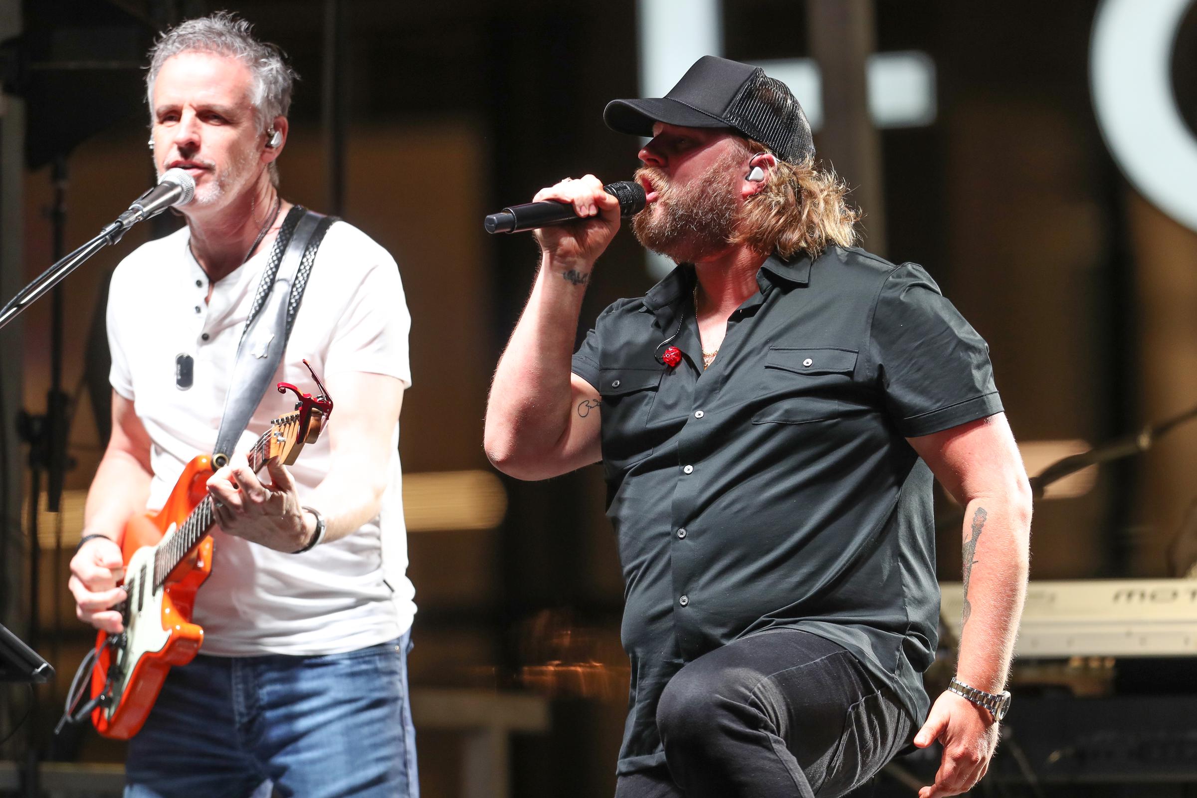 Nate Smith powers through an energetic performance with one knee raised and a microphone in hand, singing beside a guitarist as his tattooed arms peek from a fitted black shirt.