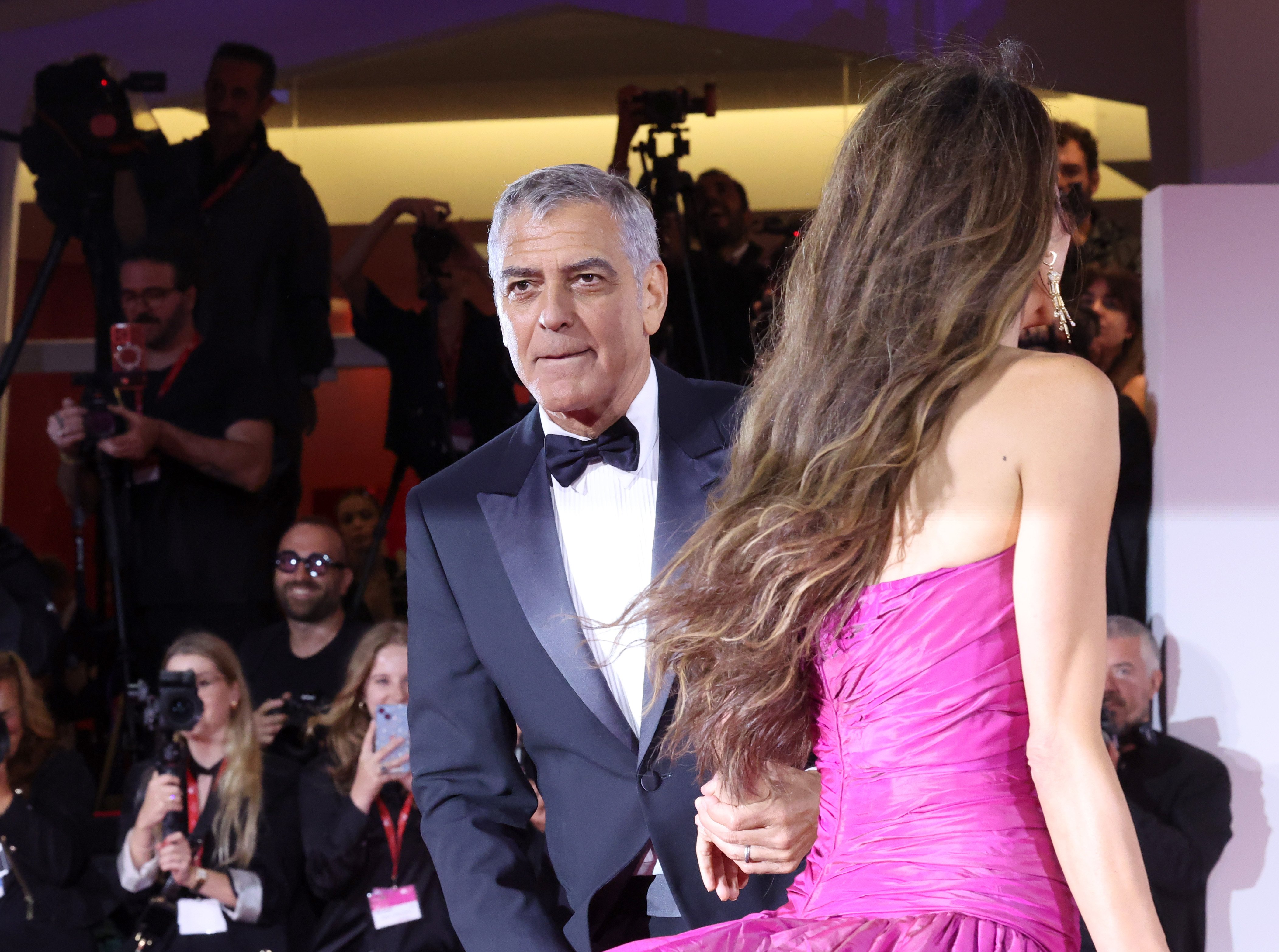 George and Amal Clooney attend the "Jay Kelly" red carpet during the 82nd Venice International Film Festival on August 28, 2025, in Venice, Italy. The actor clasps his wife's hand while gazing ahead, dressed in a sharp tuxedo, as Amal, seen from behind, showcases her voluminous waves and fuchsia vintage gown.