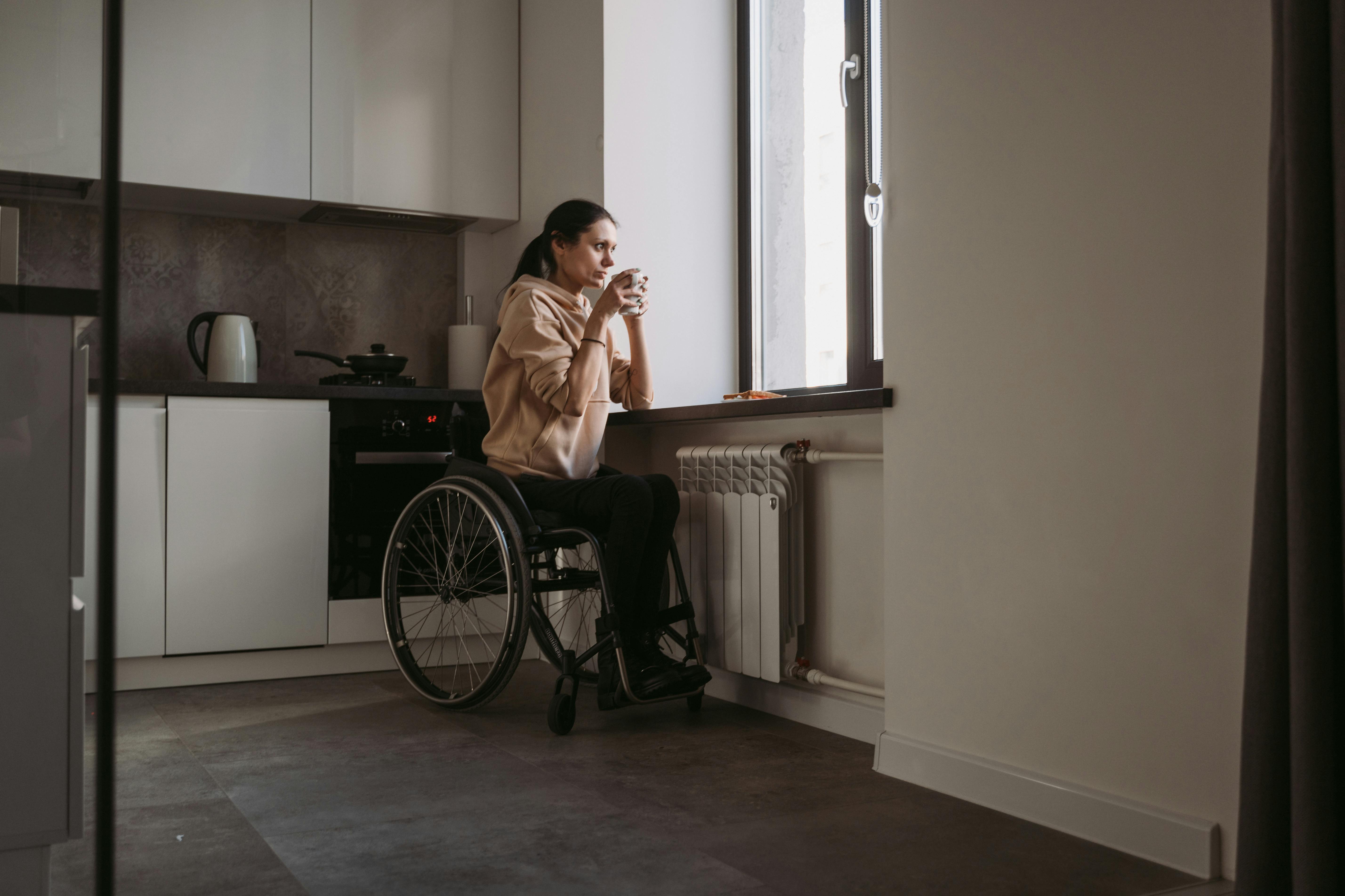 A young woman in a wheelchair drinking coffee while staring out the window in thought. For illustration purposes only | Source: Pexels