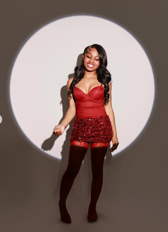 Patience Price poses confidently under a circular spotlight, wearing a coordinated red outfit with sheer tights, her long styled hair framing her face as she smiles softly in a polished studio setting. | Source: Instagram/lilpaypay.__'s profile picture lilpaypay.__