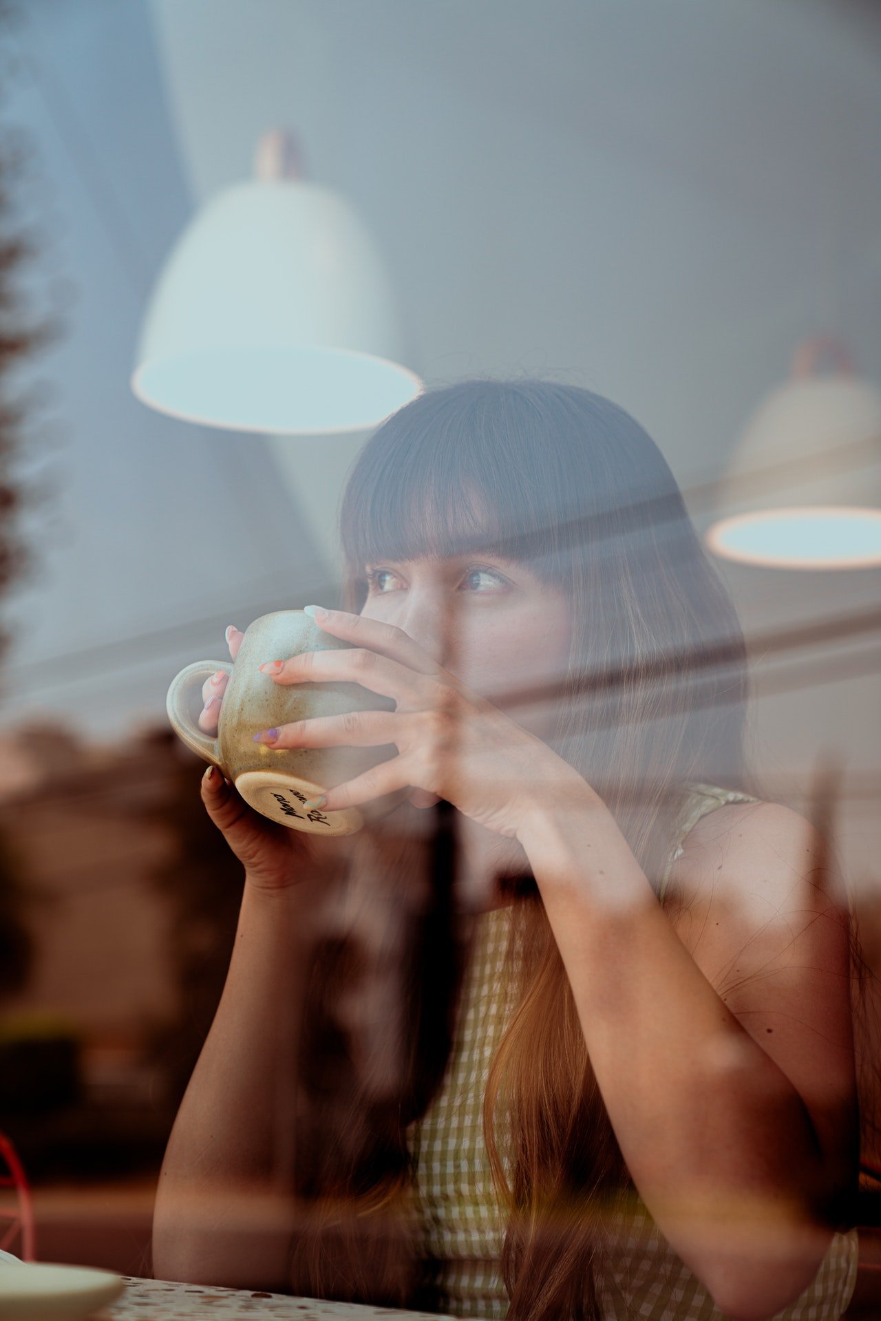 Una mujer mira a través de la ventana mientras toma una bebida de una gran taza. | Foto: Pexels
