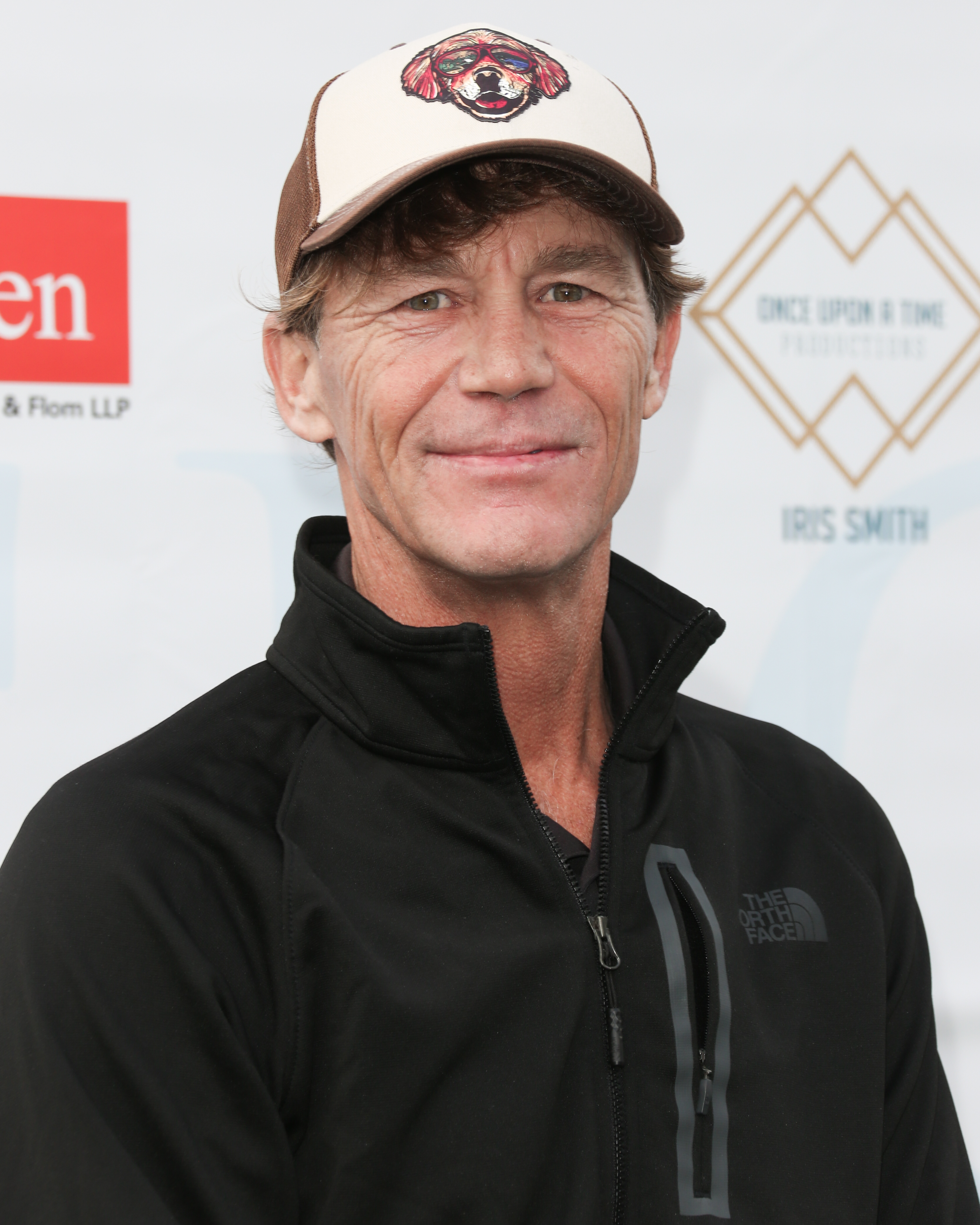 Brian Krause is seen at a public event in 2021, dressed casually in a black North Face jacket and a graphic baseball cap. Smiling softly for the camera, he stands against a step-and-repeat backdrop, giving the portrait a relaxed, approachable tone.