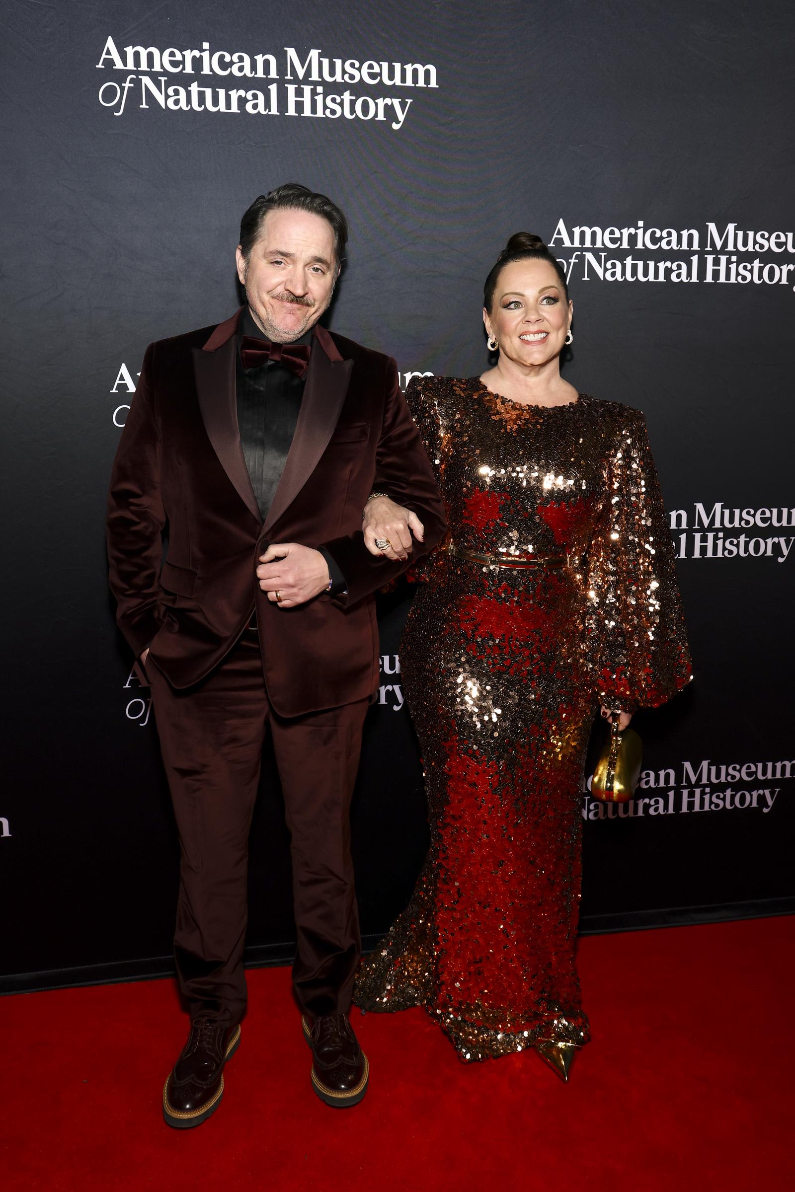 Ben Falcone stands sharp in a deep burgundy suit while Melissa McCarthy dazzles beside him in a fiery sequin gown that glimmers against the gala lights. Arm in arm, they bring a warm, charismatic presence to the red carpet, perfectly complementing each other's style.