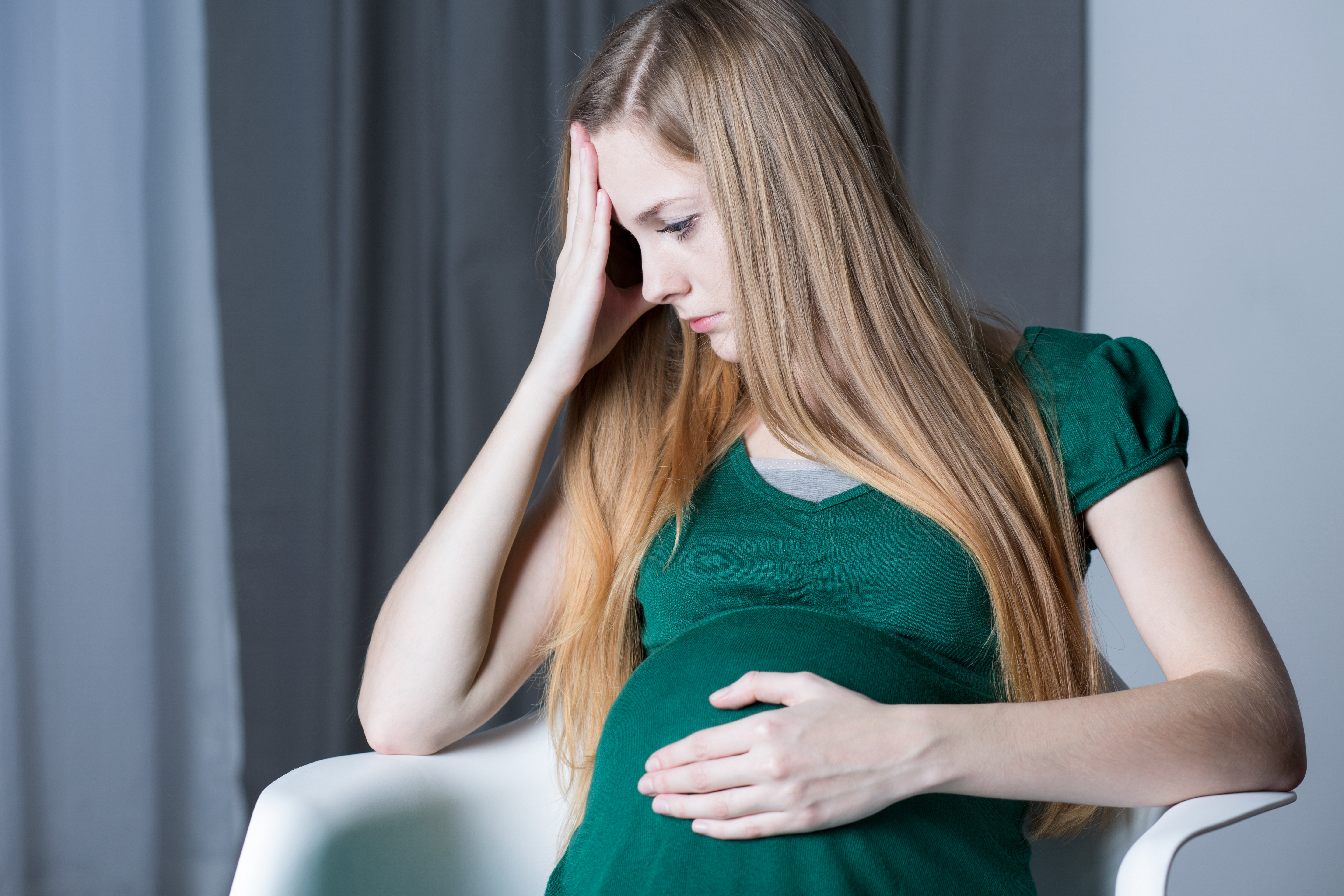 An upset pregnant woman sitting an holding her head and belly | Source: Shutterstock