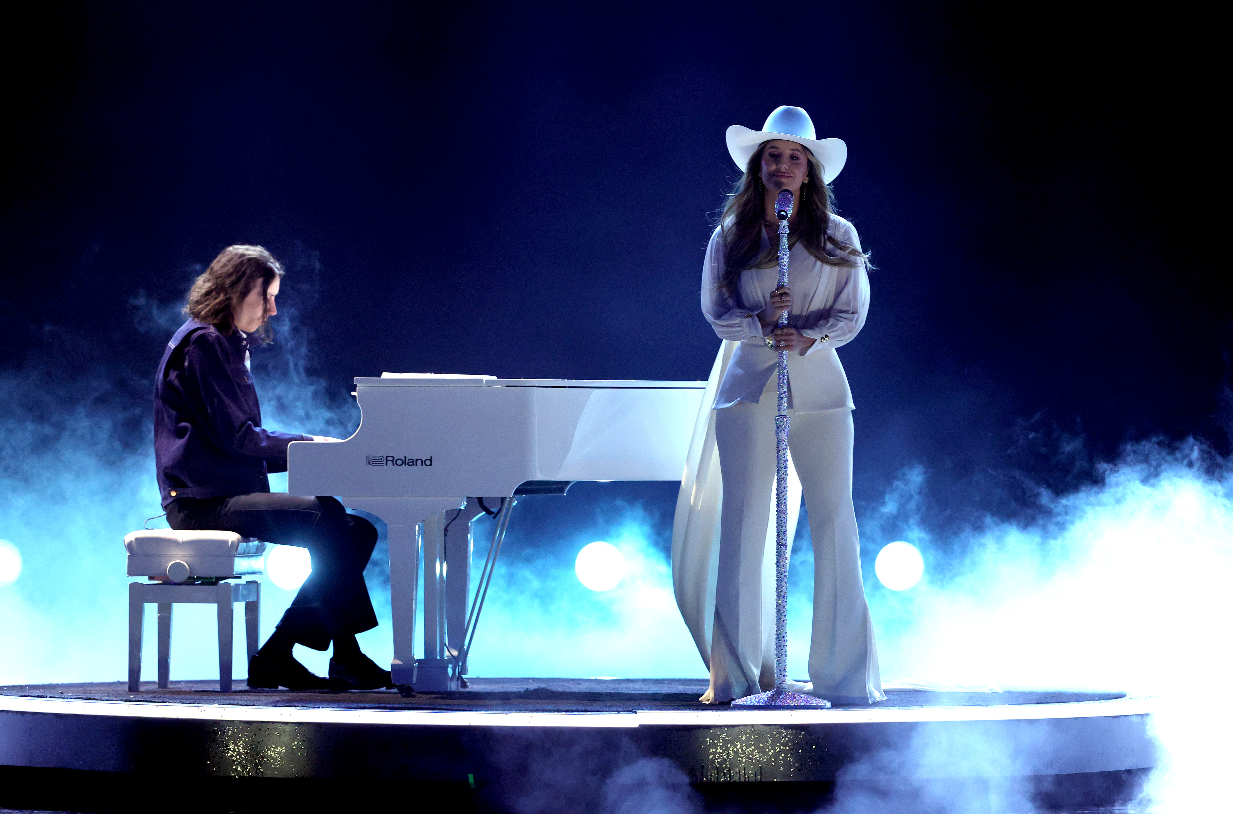 The scene is undeniably beautiful as Wilson sings beside her pianist, stage lights casting a warm glow through gentle waves of stage smoke. The soft haze curling around her feet adds a dreamlike quality, making the moment feel cinematic.