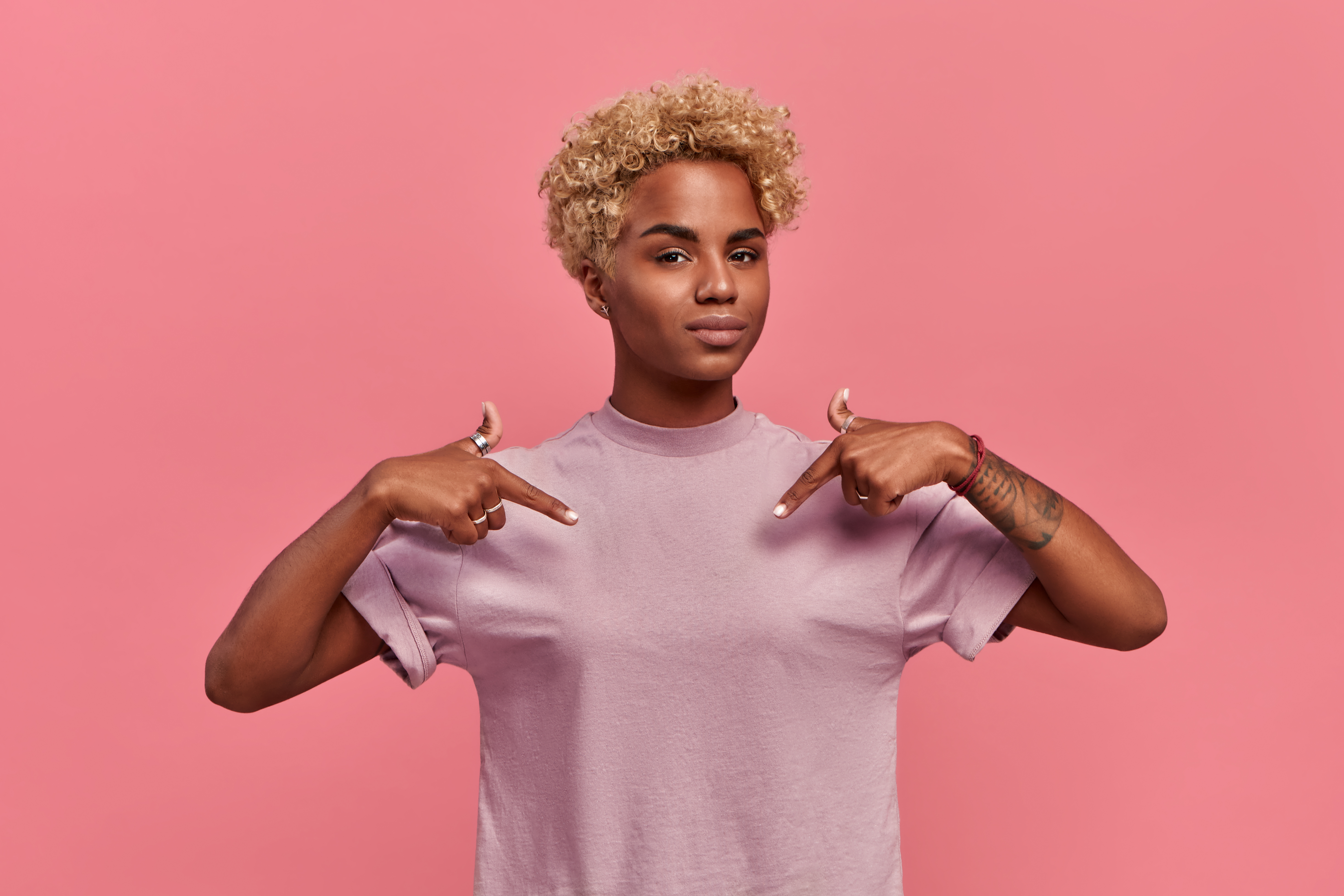 Self assured proud African American woman has blonde hairstyle satisfied with her own high achievements, points with two hands at herself, or at a t-shirt for printing, isolated over pink background | Source: Getty Images