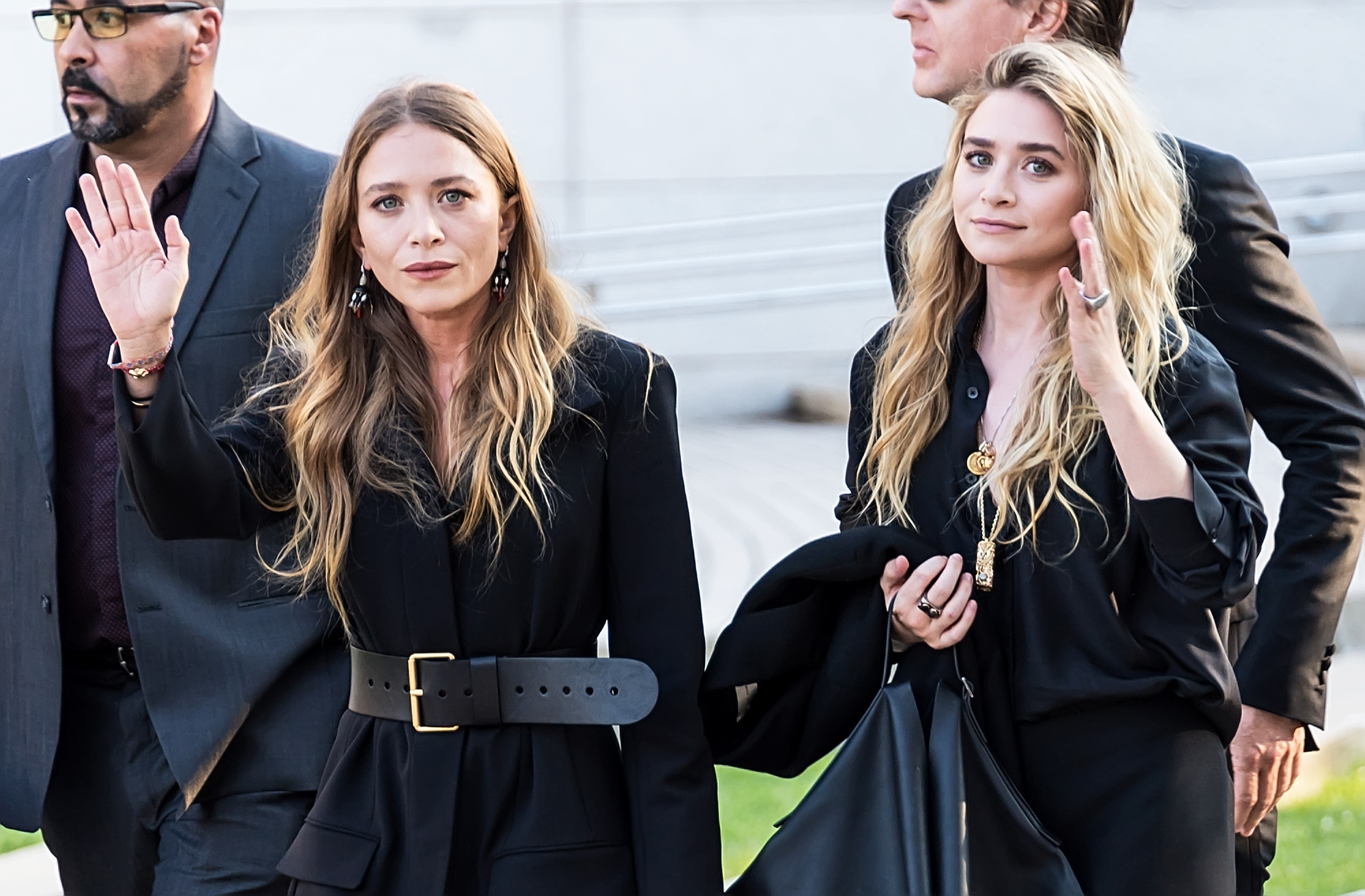 Mary-Kate and Ashley Olsen walk side by side at an event, both in structured black outfits. One raises a hand mid-gesture, while the other looks on with a calm, composed expression.