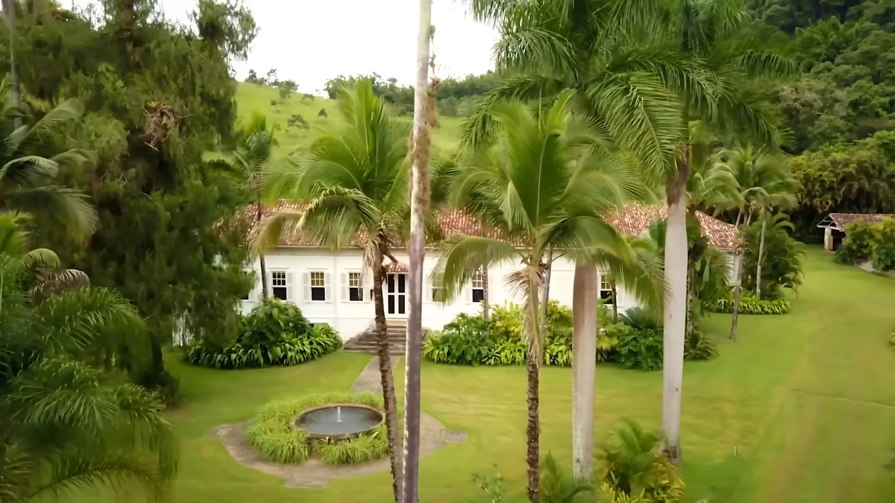 An aerial view of the main 19th-century Portuguese colonial-style farmhouse, nestled in a lush, mountainous valley and surrounded by towering palm trees. | Source: YouTube/@Archdigest