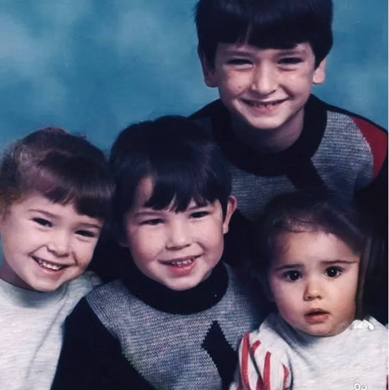 An old Tweedy family portrait shows four children pressed together against a powder-blue studio backdrop, each one mid-smile for the camera. The eldest, in a grey and red jumper, stands at the back with his arms around the younger three, who range from a grinning toddler in a red-striped top to two slightly older children, one wearing a matching patterned knit. The photograph was shared to TikTok on 29 August 2021. | Source: TikTok/__lottiehulmeshowbiz