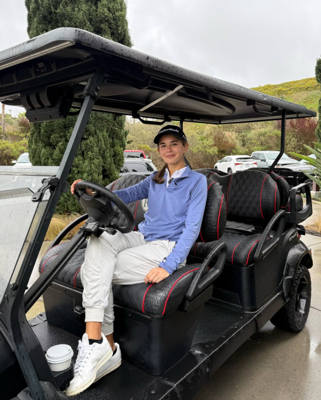 Kai sits behind the wheel of a golf cart, dressed in a blue top and white pants, smiling with a coffee cup nearby | Source: Facebook/Kai-Trump