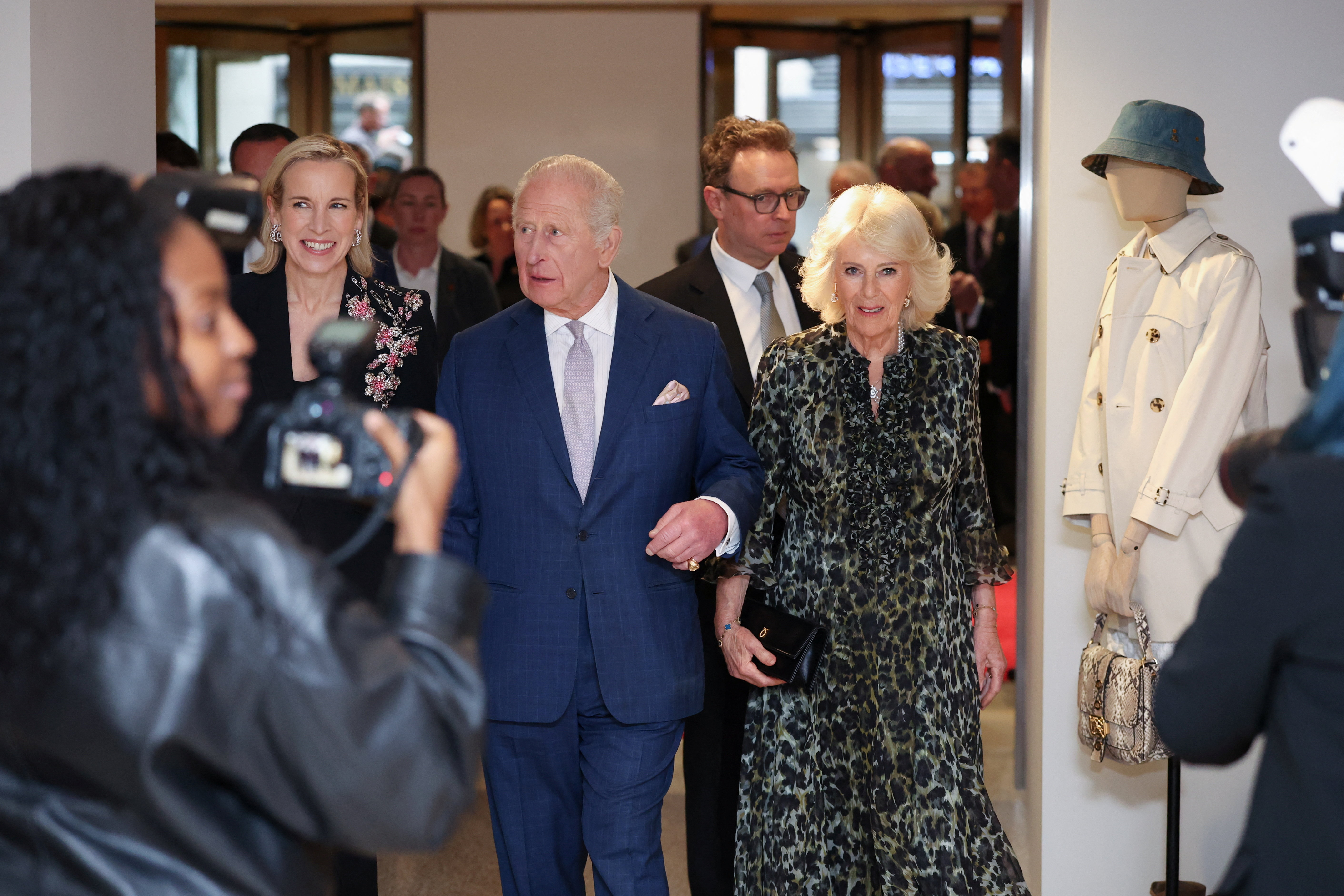 King Charles III, dressed in a classic navy blue suit with a pale lavender tie and white pocket square, escorts Queen Camilla — elegant in a dark olive and black leopard-print dress with a small black clutch — as the royal couple makes their entrance into the venue, cameras rolling and attendees beaming behind them.