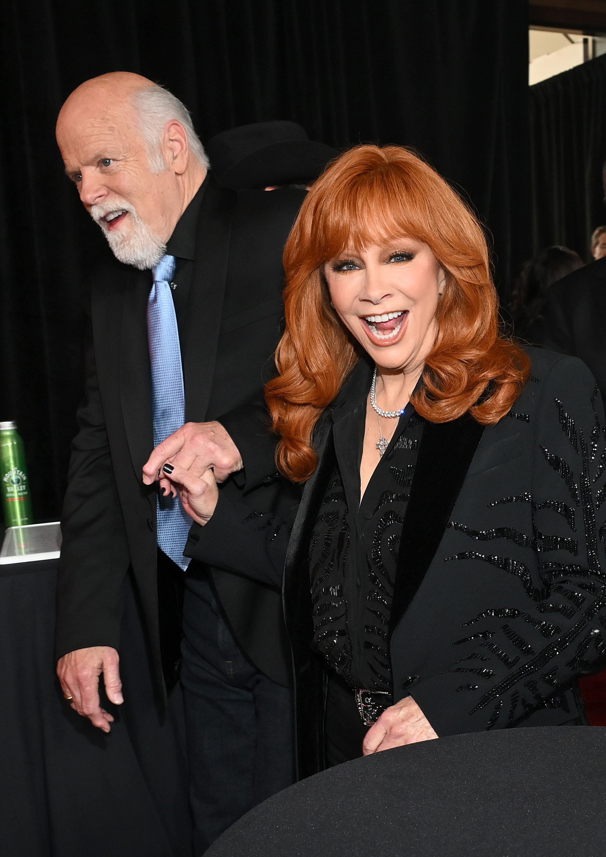 Reba McEntire flashes a radiant smile in a beaded black suit, holding hands with Rex Linn as they make their way through the crowd with playful energy