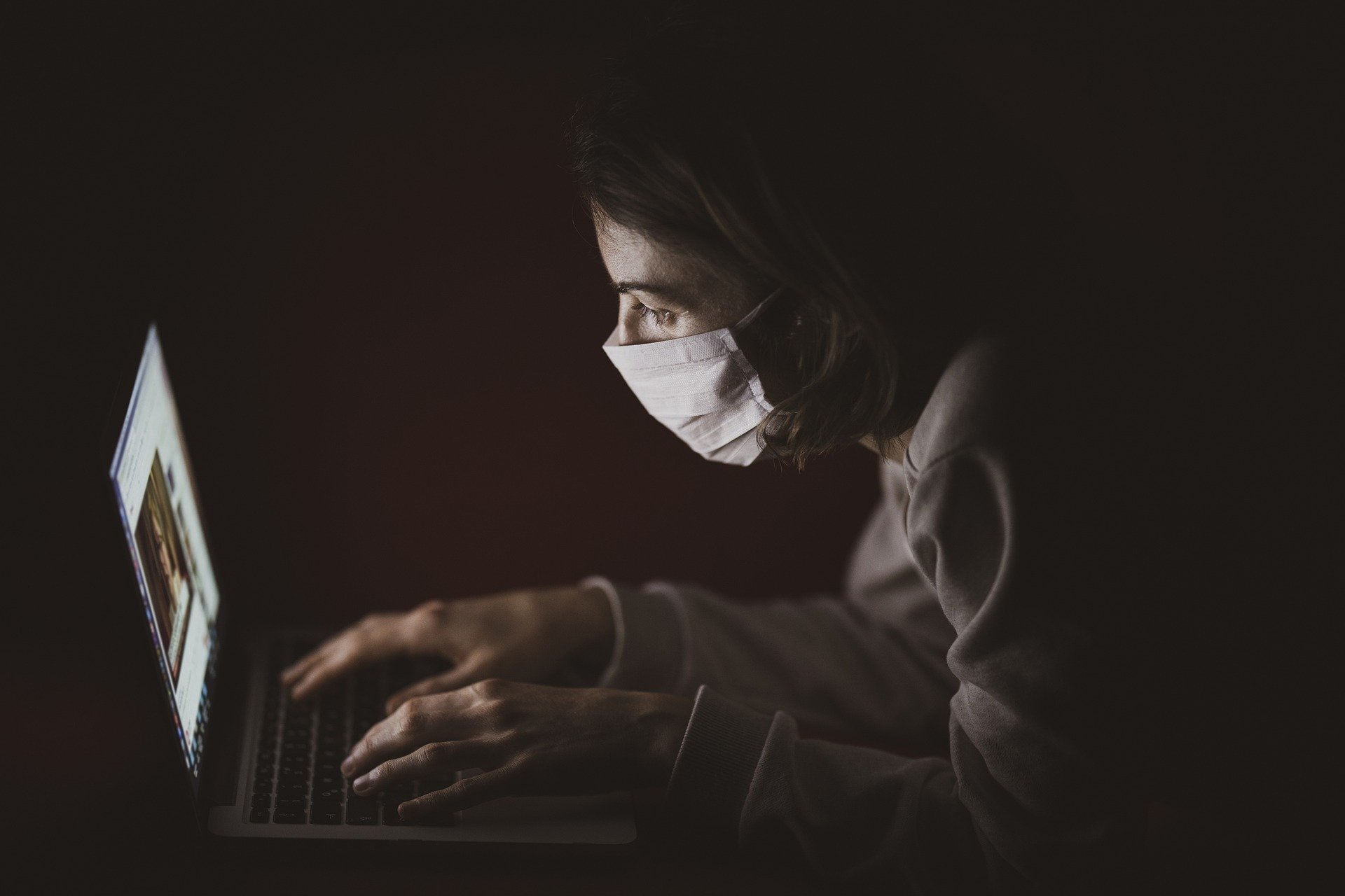 A man sitting at his laptop in the dark wearing a face mask amid the novel coronavirus pandemic. | Source: Pixabay.
