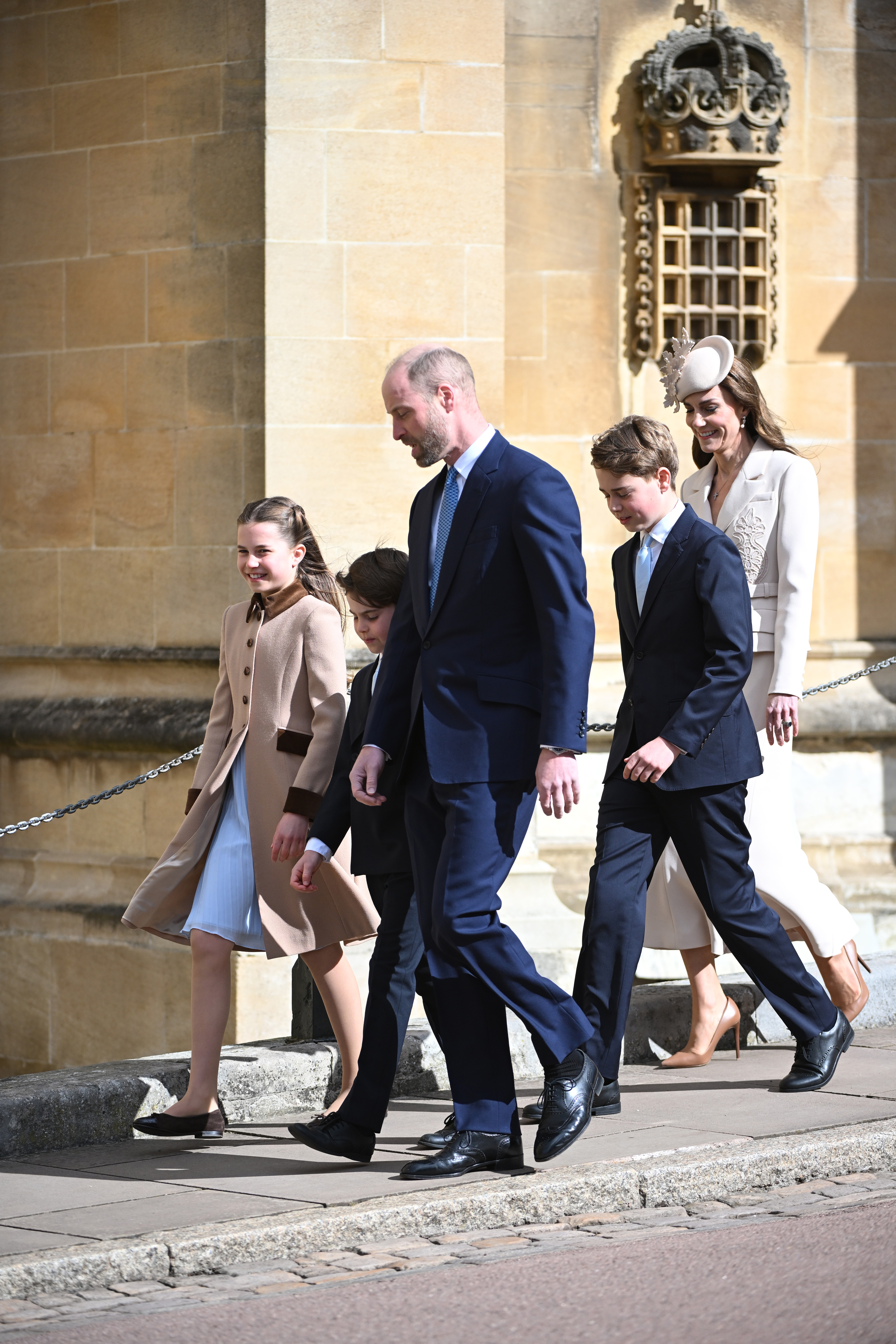 The family stepping up towards the chapel entrance, the ornate stone crown carved into the pillar above them. Princess Charlotte laughs as she walks on the left, Prince Louis and the Prince of Wales stride ahead in the centre, and Prince George walks alongside the Princess of Wales just behind them, the lace appliqué detail of her blazer catching the bright morning light.