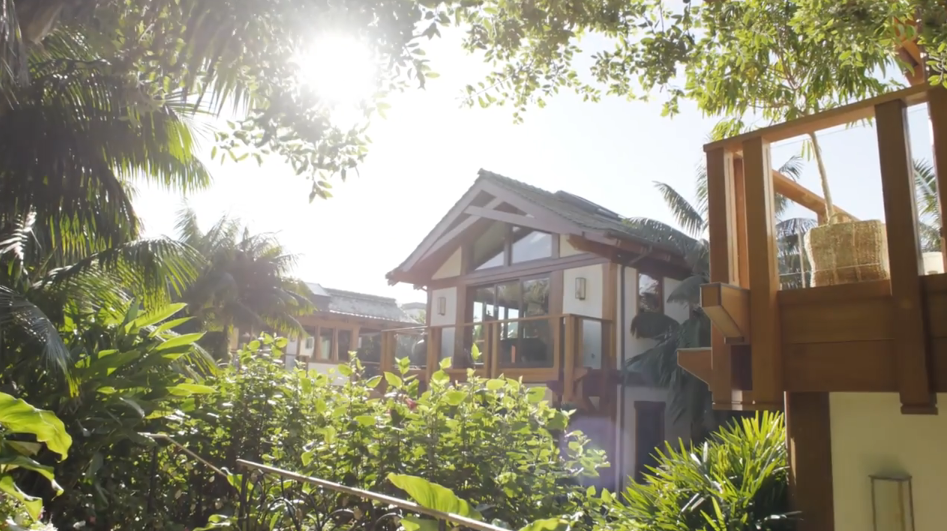 Sunlight filters through a dense canopy of tropical trees and palm fronds, highlighting the warm timber gables and second-story balcony of the residential pavilion. Lush green garden beds and wooden architectural detailing surround the property in a secluded, resort-like atmosphere. | Source: YouTube/Architectural Digest