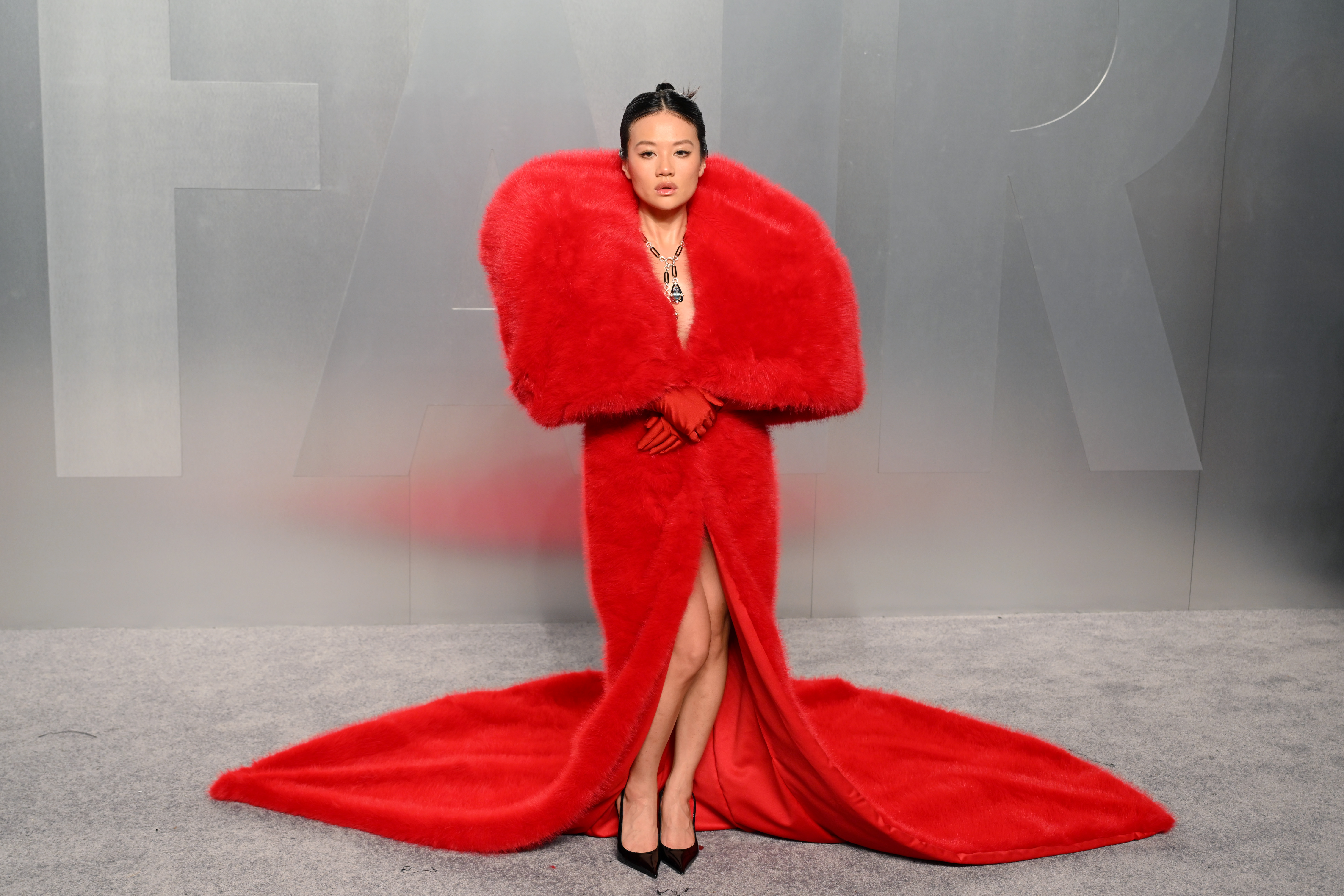 Rei Ami arrives fully enveloped in a floor-length red faux fur coat with an enormous exaggerated collar that engulfs the shoulders and neck, a deep V-neckline revealing a statement necklace, a front slit exposing the legs, red gloves, black pointed-toe heels and a dramatic sweeping train.