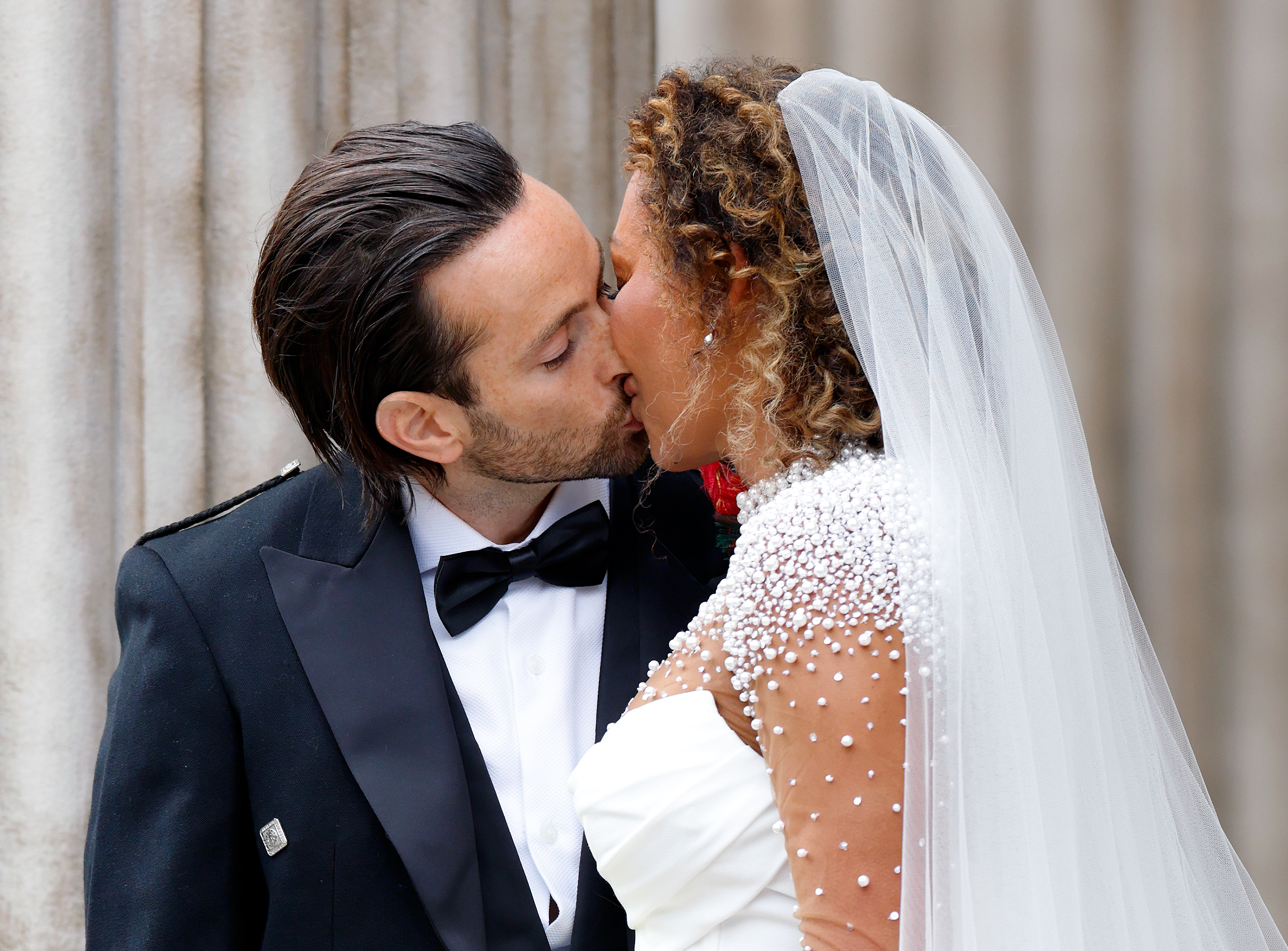 A close-up captures the couple locked in a passionate kiss, their faces framed in a moment of pure emotion. Mel B’s eyes are gently closed while McPhee leans in, matching her energy with quiet intensity.