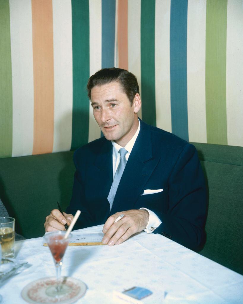 Australian actor, sitting on a green seat and sitting at a table, on which drinks rest, with striped wallpaper in the background, 1950.  | Photo: Getty Images