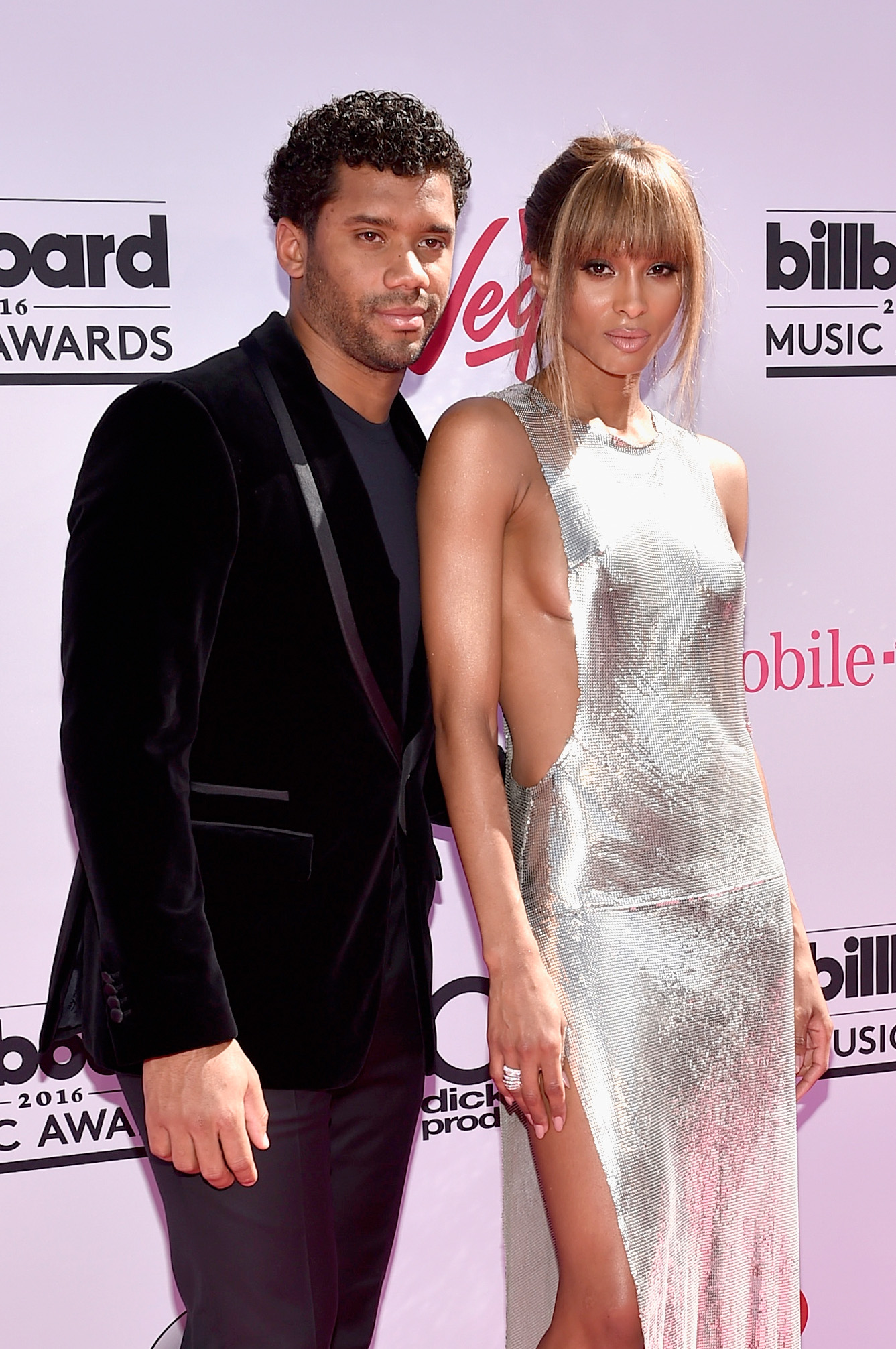 Ciara commands attention in a shimmering silver dress with a high slit, standing confidently next to Russell Wilson on the red carpet. He maintains a composed stance beside her, allowing her bold, eye-catching look to take center stage.