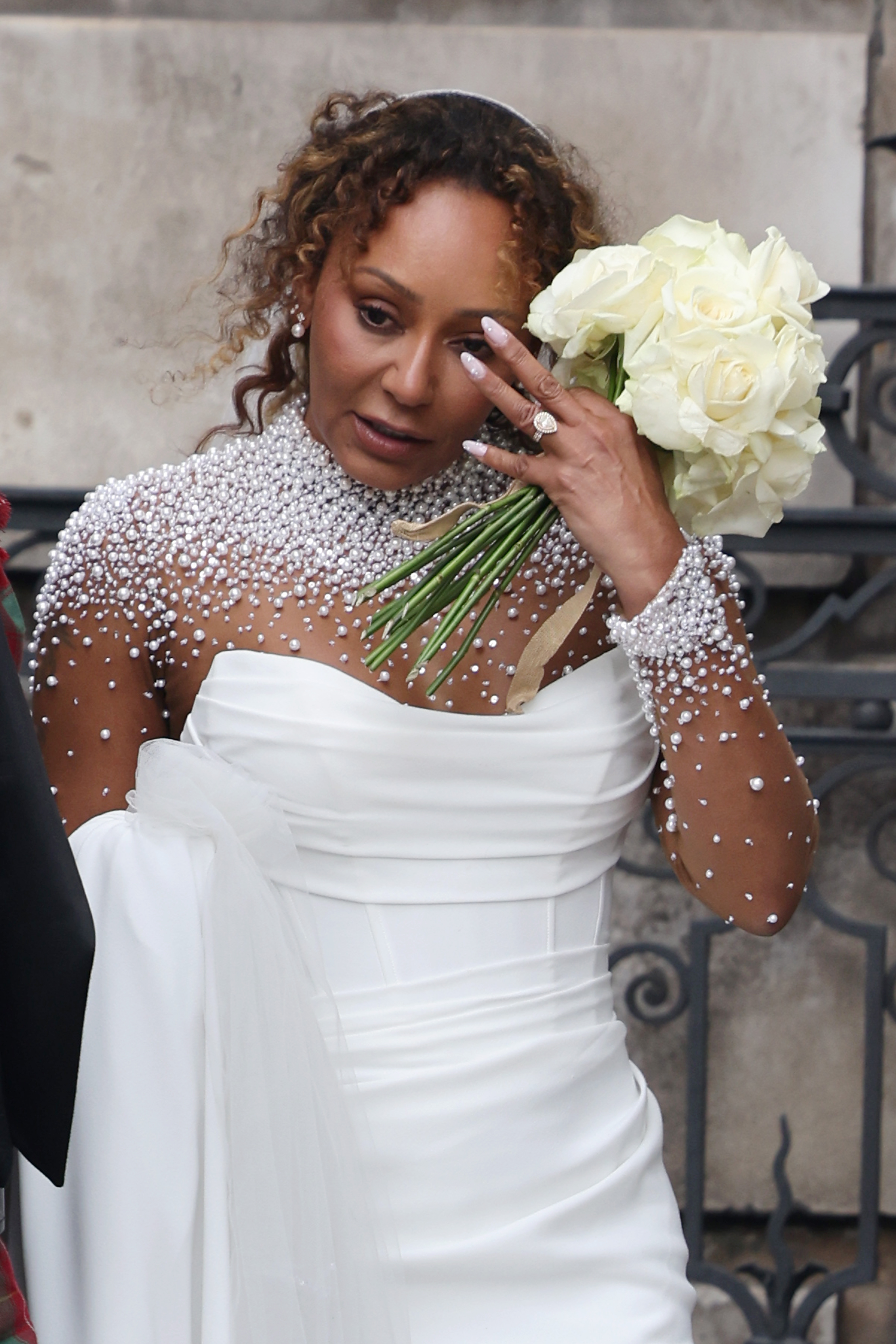 Mel B is seen wiping something from her face while holding her bouquet. Her wedding ring catches the light, clearly visible on her hand. The simple gesture adds a touch of authenticity to the celebration, highlighting the quiet, personal moments behind the public event.