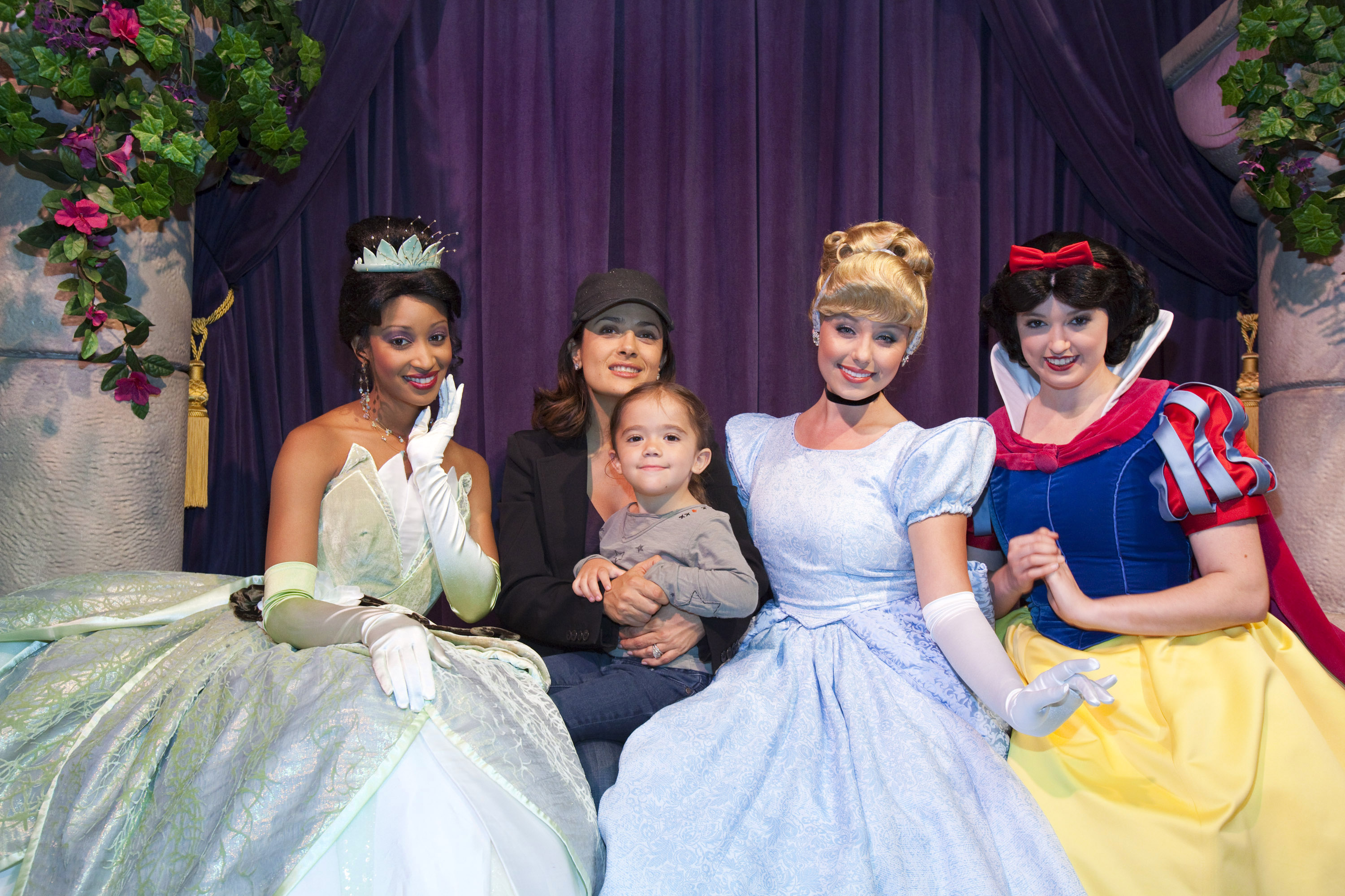 In a charming family outing to Disneyland, Salma Hayek and Valentina Pinault pose alongside iconic Disney princesses Tiana, Cinderella, and Snow White. Salma, dressed casually in a black cap and jacket, beams while holding her daughter close. Valentina sits comfortably on her mom's lap, wearing a gray top and jeans, her expression a mix of awe and quiet delight.