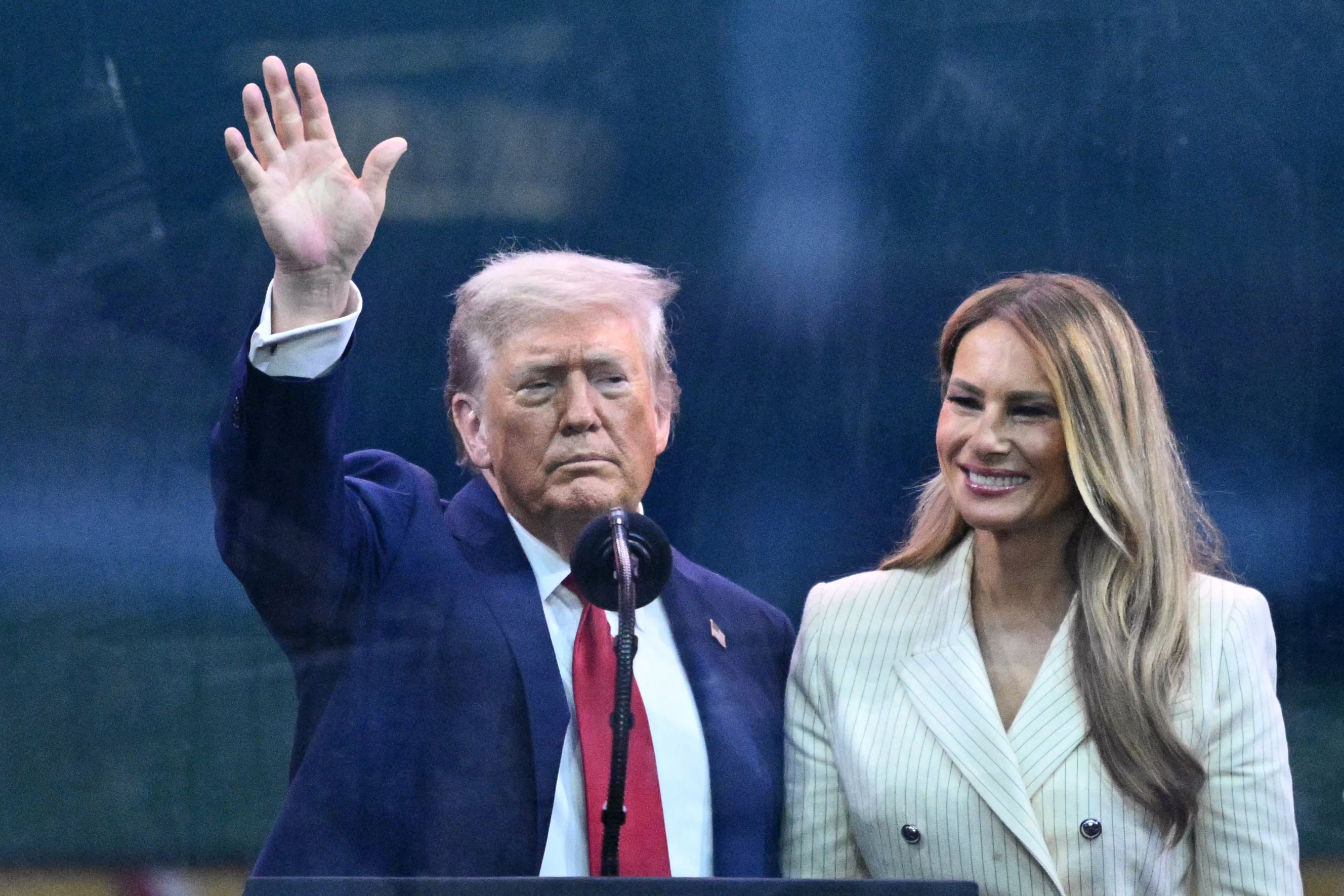 Donald Trump waves to the crowd beside Melania Trump as they attend the U.S. Army’s 250th anniversary parade on June 14, 2025. Standing behind glass, Melania smiles in a pinstripe outfit while Trump acknowledges attendees with a raised hand.