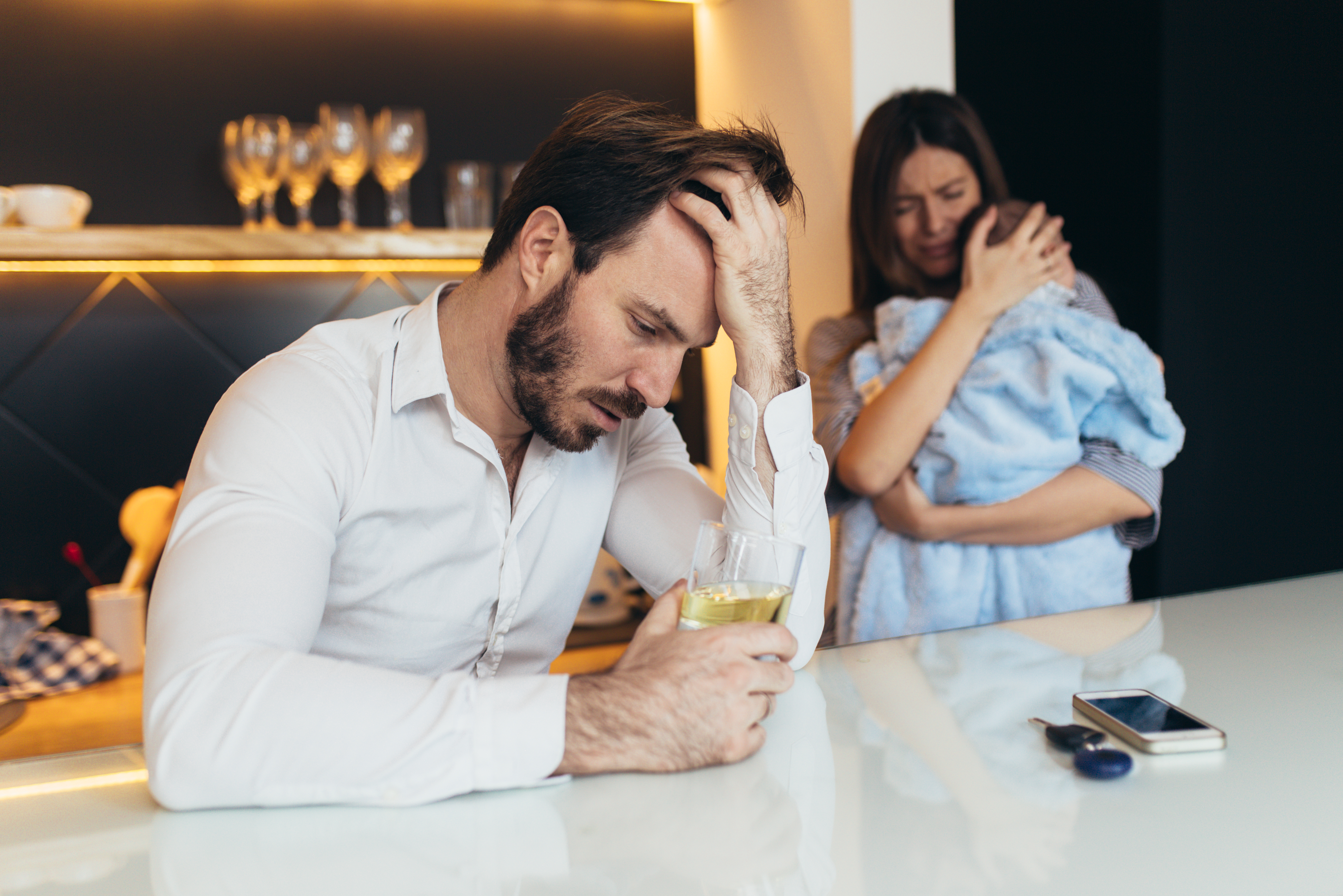An emotional woman holding a child while standing behind a depressed man who is drinking | Source: Shutterstock
