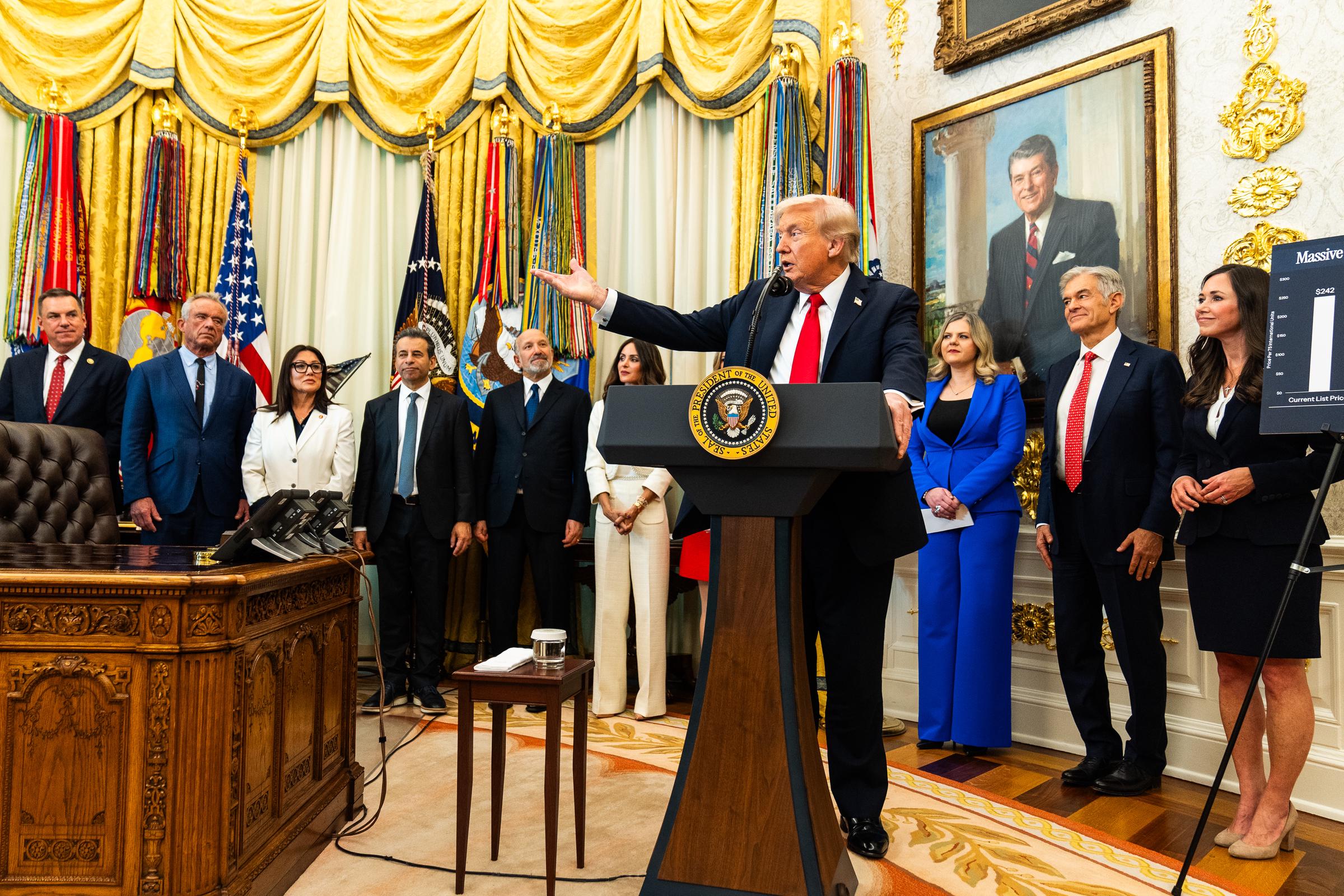 President Donald Trump speaks during an event on October 16, 2025, in the White House. The Oval Office is framed by gold-trimmed drapes, military banners, and the iconic Resolute Desk beneath a portrait-lined wall.