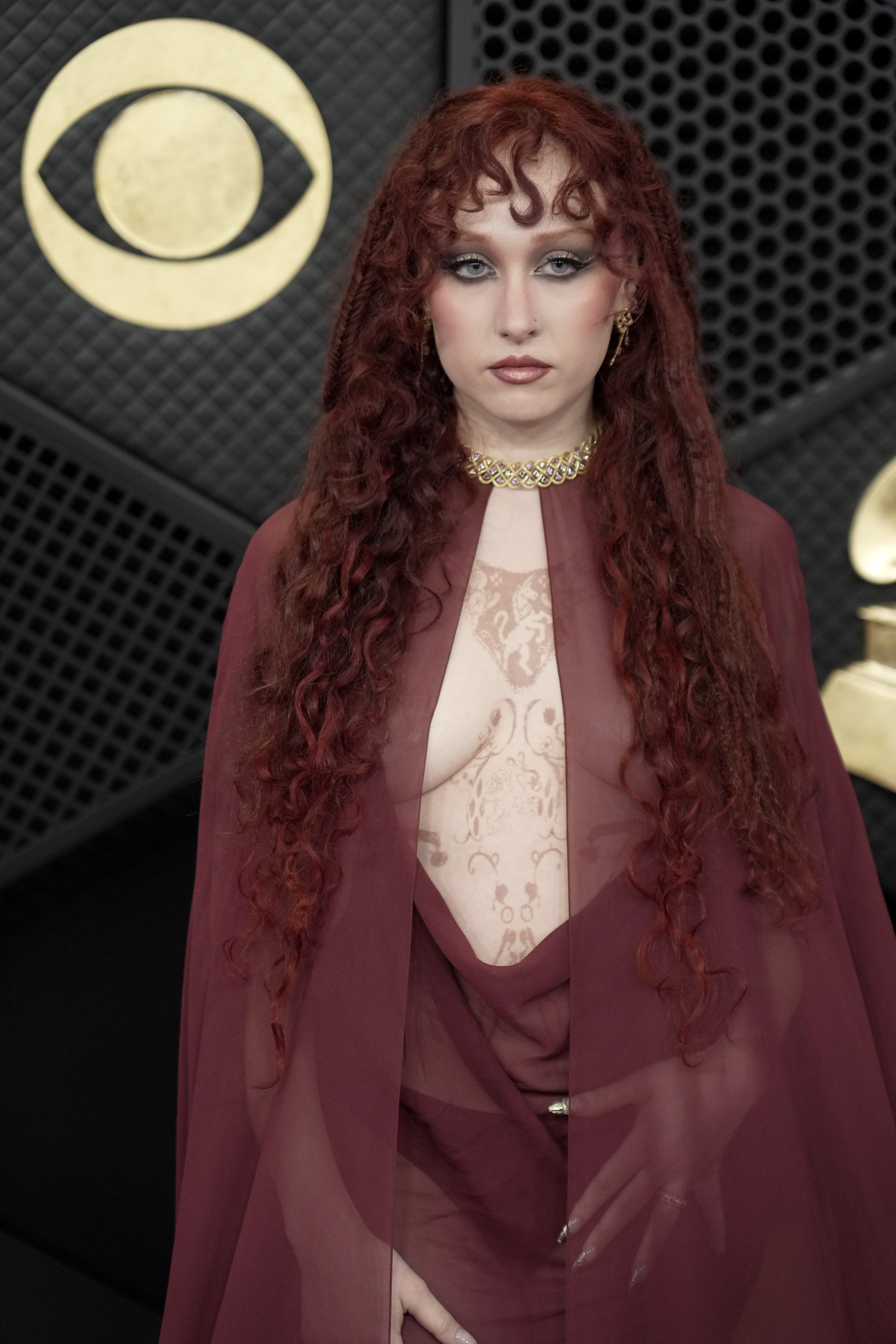 Chappell Roan is photographed on the red carpet at the 68th Grammy Awards at Crypto.com Arena in Los Angeles, California, wearing a sheer, deep red ensemble layered with a flowing, cape-like piece draped over her shoulders. The translucent fabric revealed intricate body art beneath, while her voluminous curly red hair and gold choker created a striking contrast against the darker tones of the look.