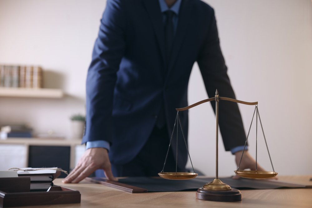 A photo of a male lawyer by table in office standing behind the scales of justice. | Photo: Shutterstock