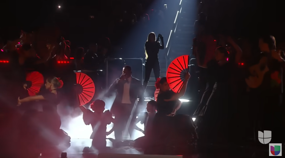 Will Smith and India Martínez stand close together on a fog-filled stage, surrounded by red lighting and flamenco-inspired dancers | Source: YouTube/univision