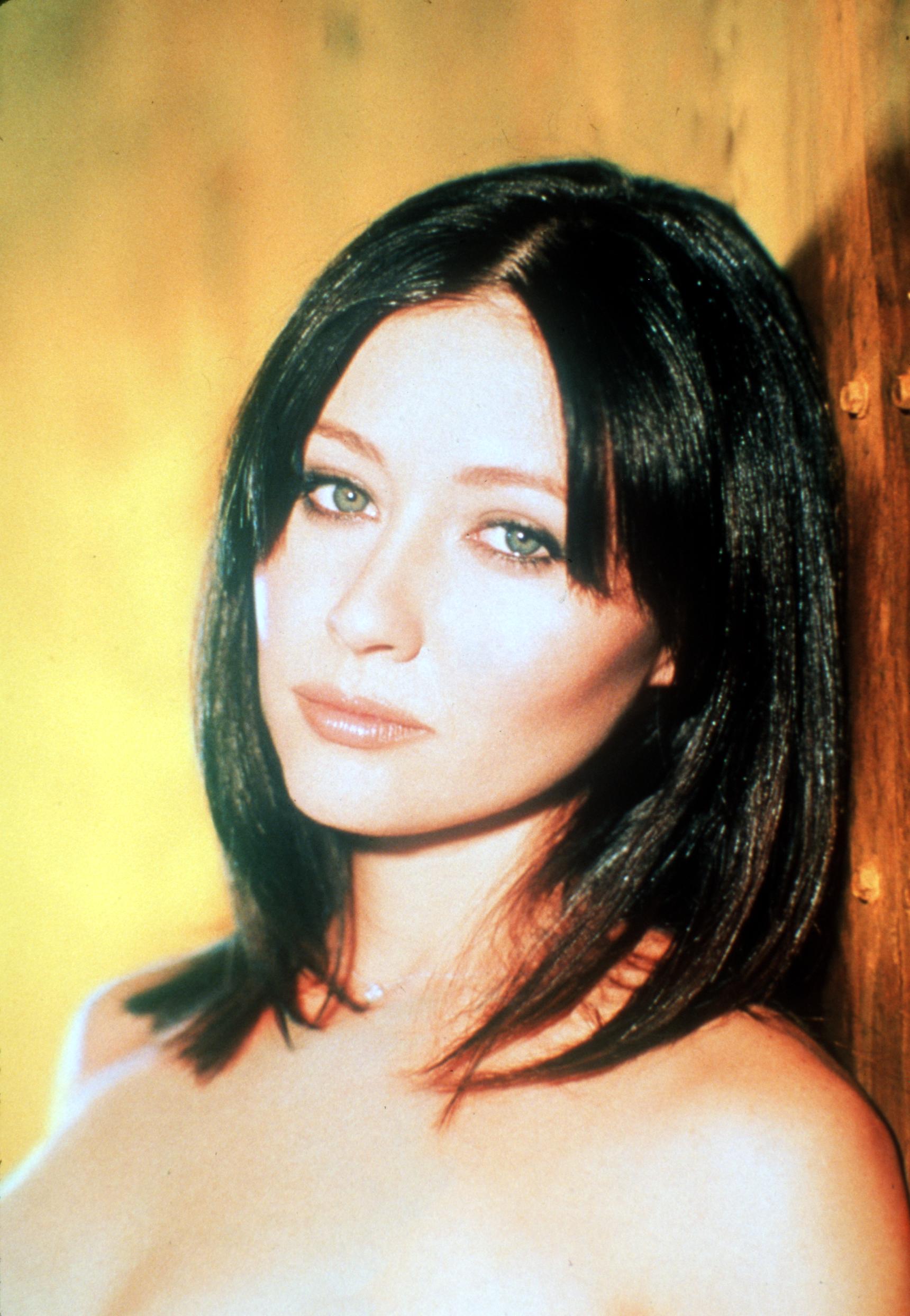 The portrait captures Shannen Doherty, who was starring in “Charmed” at the time, with straight, dark hair and a calm, introspective gaze. Set against a warm, golden background, her light eyes and softly lit features create a quiet, intimate mood. A delicate necklace rests at her neck, adding a subtle detail to the minimalist composition.