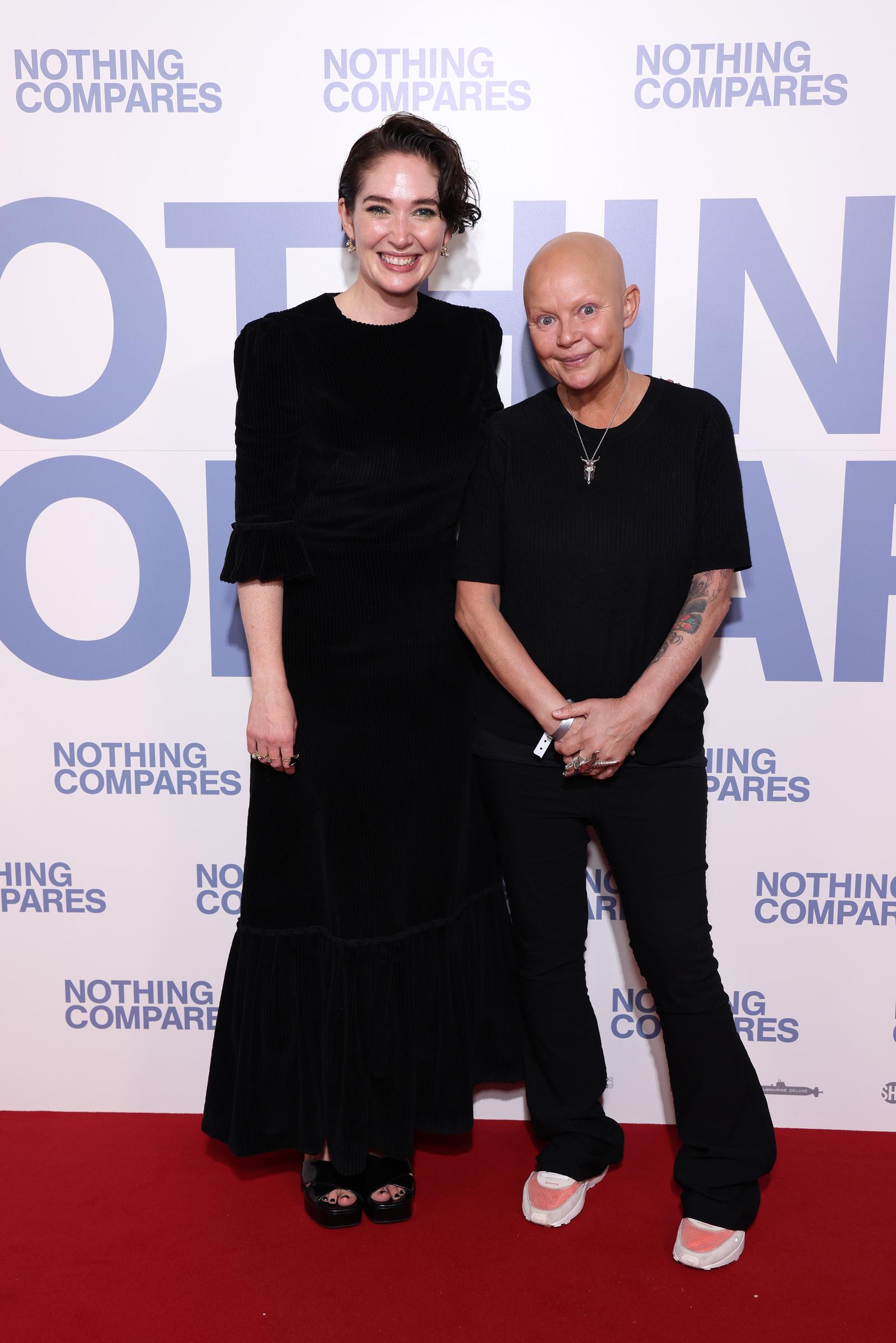 Porter stands on a red carpet beside Director Kathryn Ferguson, who wore in a long black dress, both smiling for the camera. Porter wears a plain black short-sleeved top and trousers, a cross pendant necklace, and a wristband, the "Nothing Compares" logo repeating across the backdrop behind them. Pictured at a special screening of "Nothing Compares" at Picturehouse Central, London, October 2022.