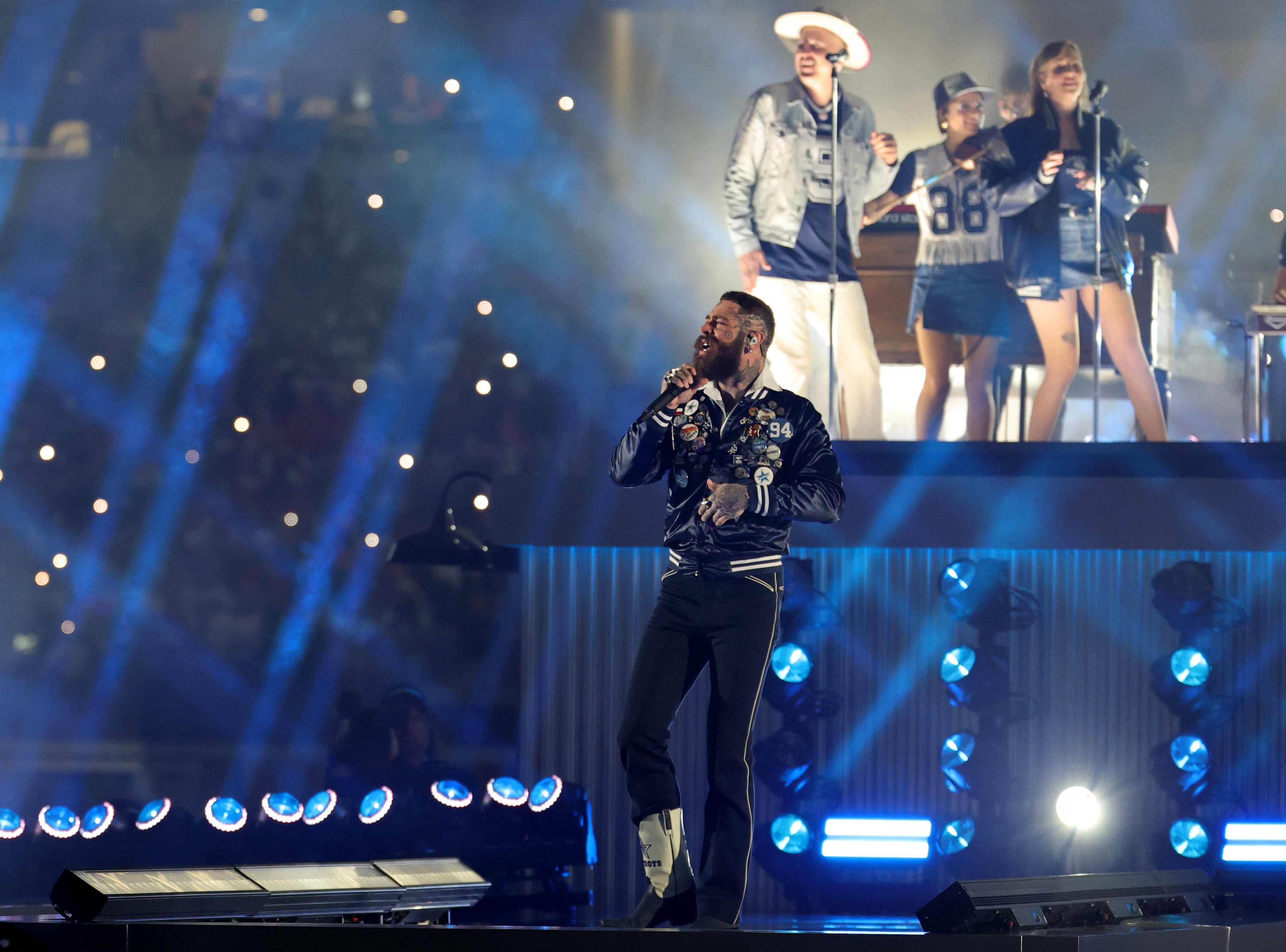 Bathed in blue stage lights, Post Malone performs in front of background singers wearing Cowboys gear during his Thanksgiving halftime show | Source: Getty Images
