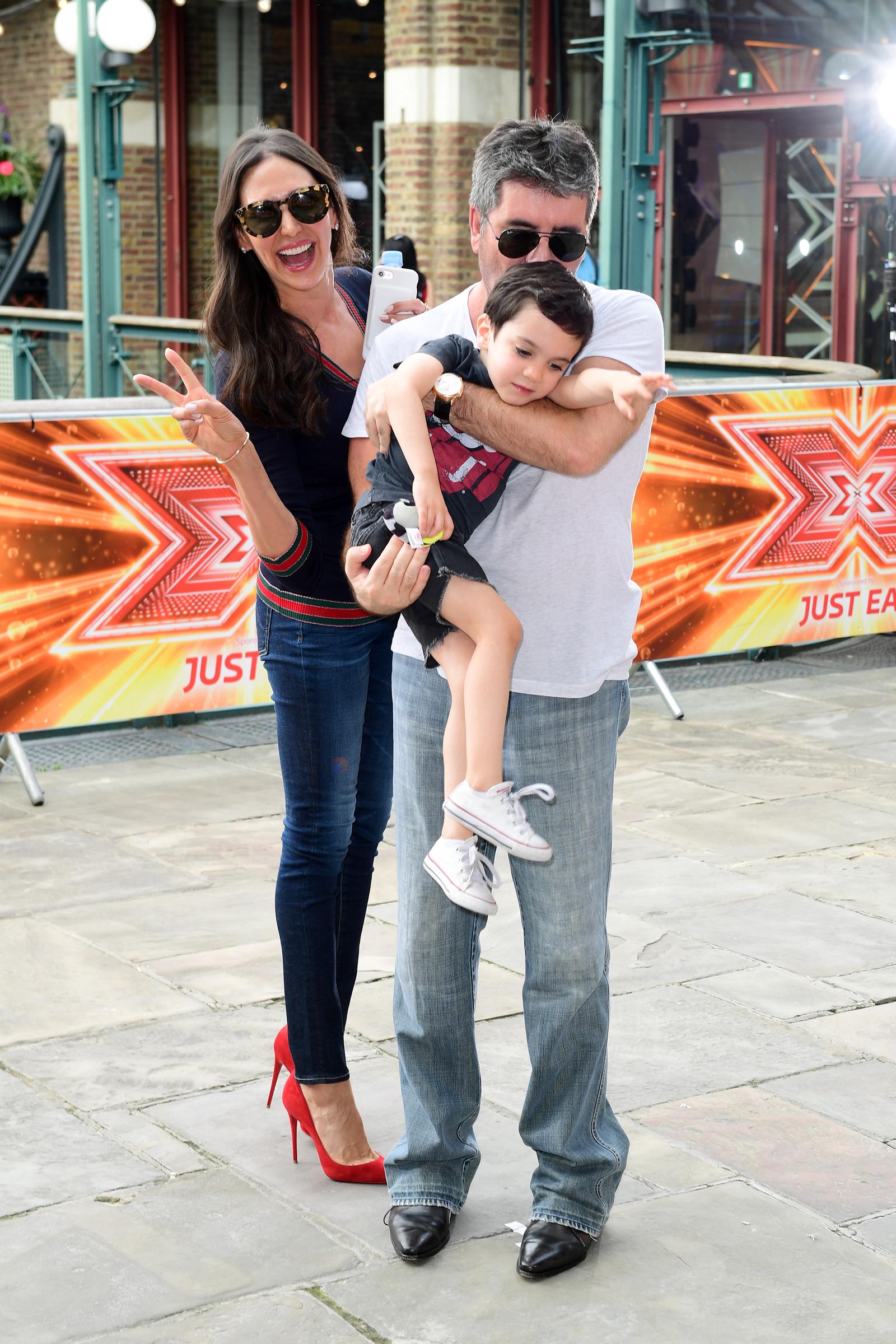 Simon Cowell, Lauren Silverman, and Eric were spotted at "The X Factor" filming at Tobacco Dock on Wapping Lane in London. The photo captures their playful side, Silverman flashes a peace sign while Cowell tries to keep hold of his restless toddler, who looks eager to wriggle free and be set down.