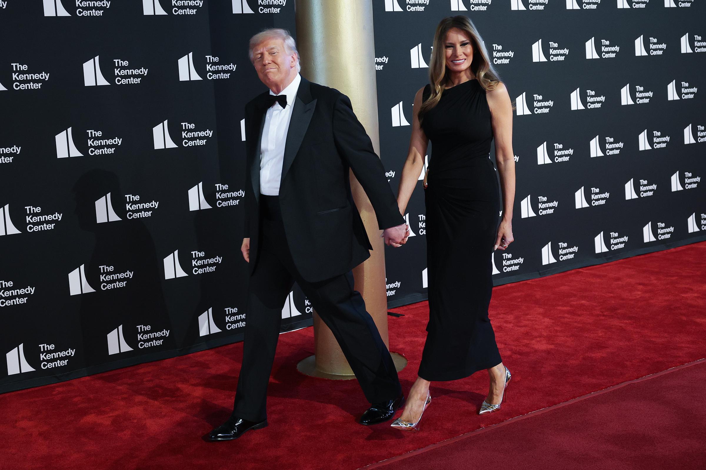 Donald and Melania walk hand in hand along the red carpet, both smiling as they make their way past the cameras. Melania’s dress shifts gently with each step, accentuating her figure.