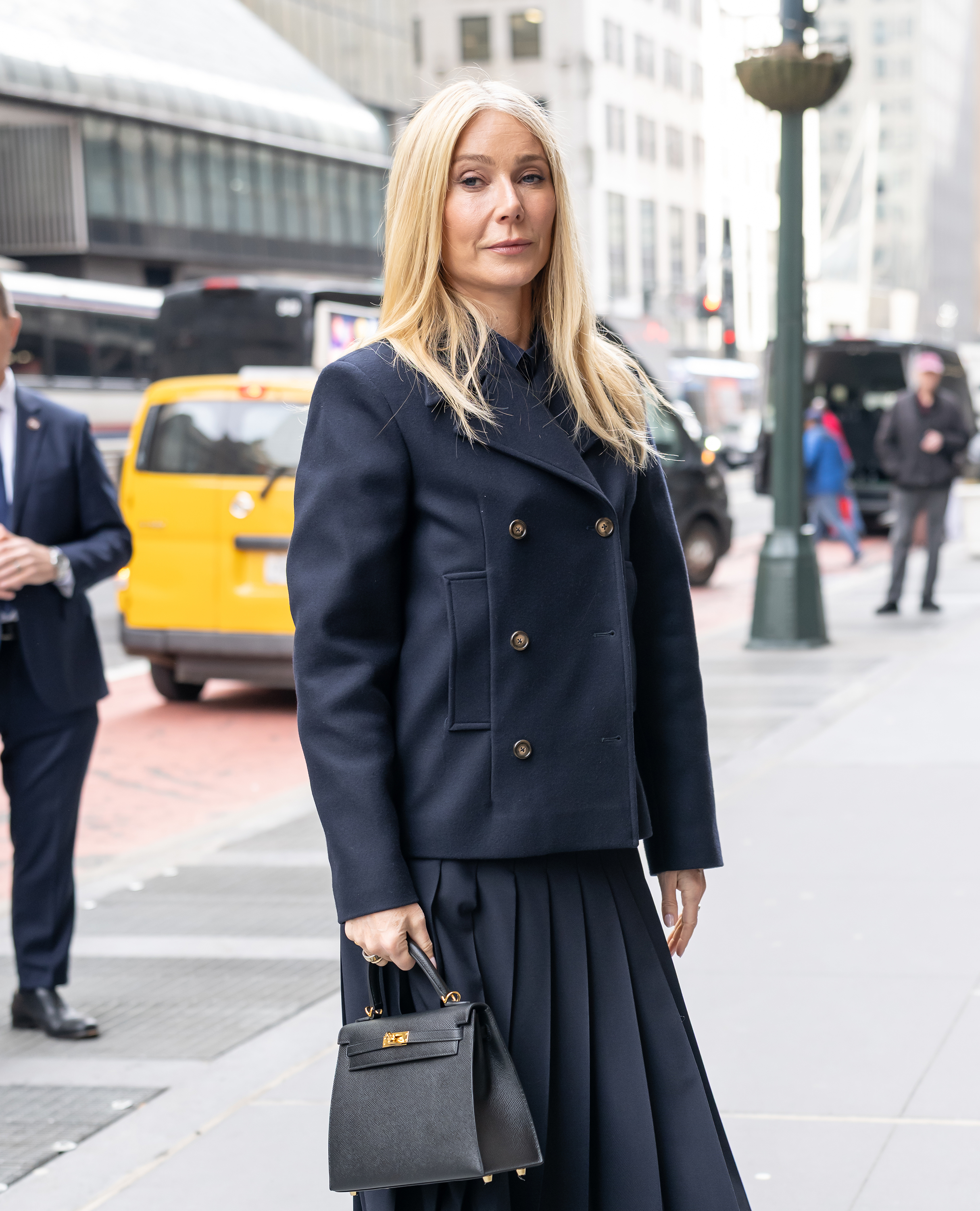 Gwyneth Paltrow stands on a New York City sidewalk, facing slightly to the side with a composed expression, her navy double-breasted jacket structured and her blonde hair worn straight.