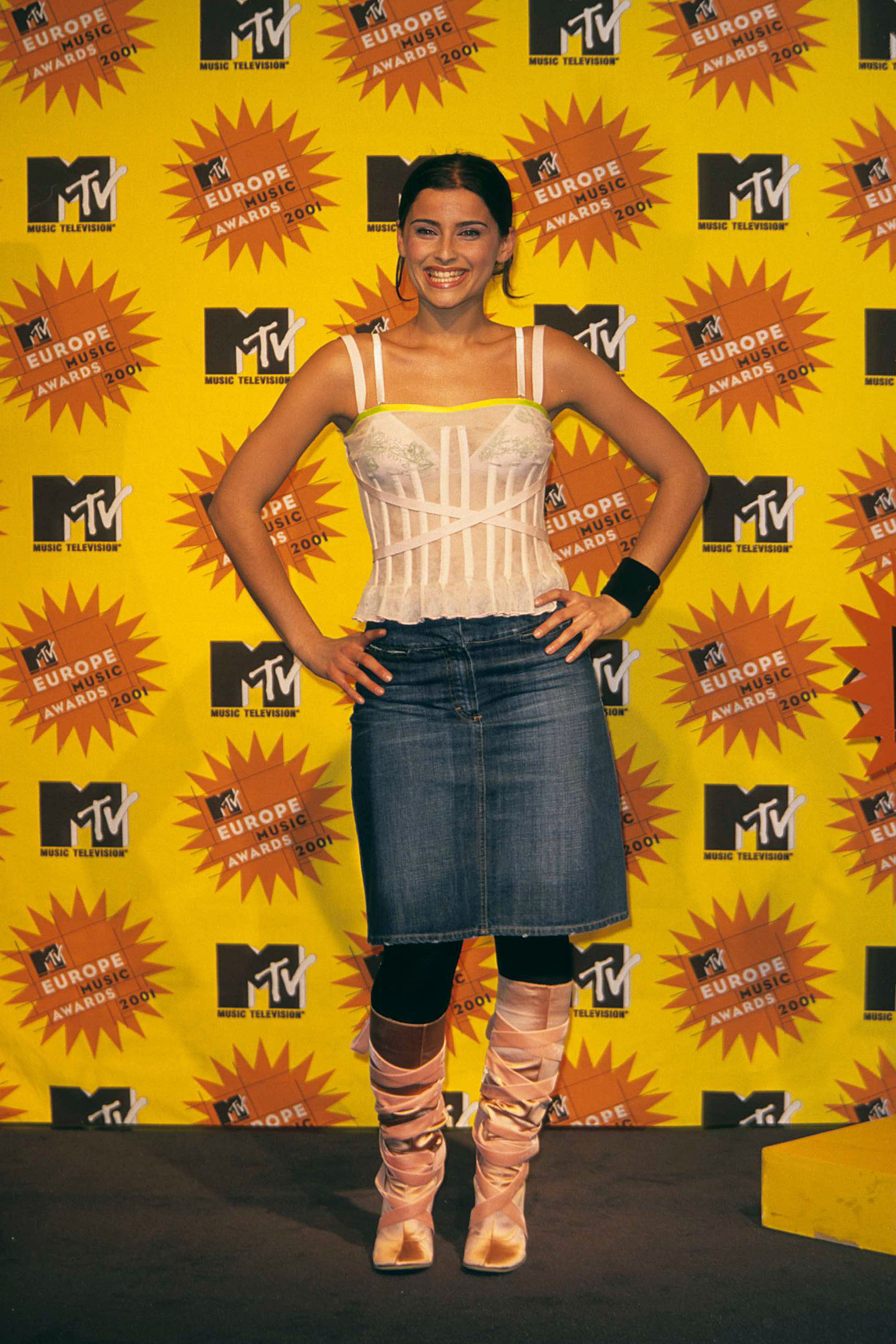 Nelly Furtado poses confidently at the MTV Europe Music Awards press room in Frankfurt, smiling in a white corset top, denim skirt, and satin boots against a bold yellow backdrop.
