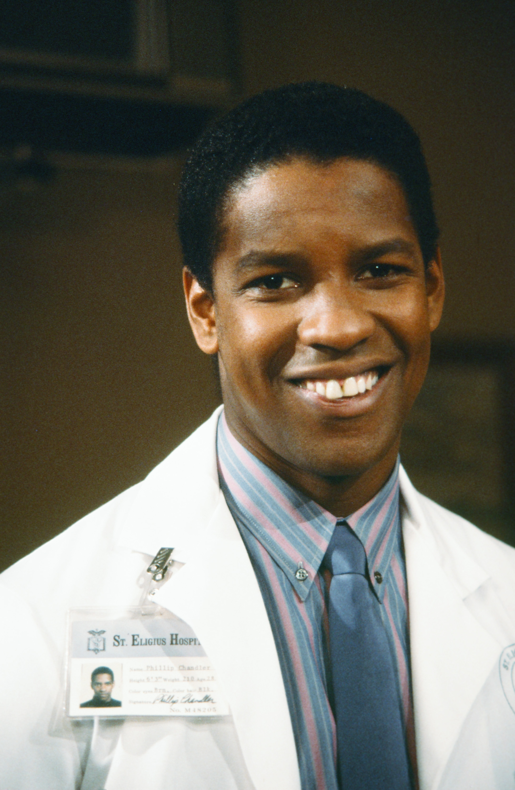 On the set of "St. Elsewhere," Denzel Washington smiles in his role as Dr. Philip Chandler. His youthful appearance and neat, natural hairstyle reflect the actor's early days in network television drama.