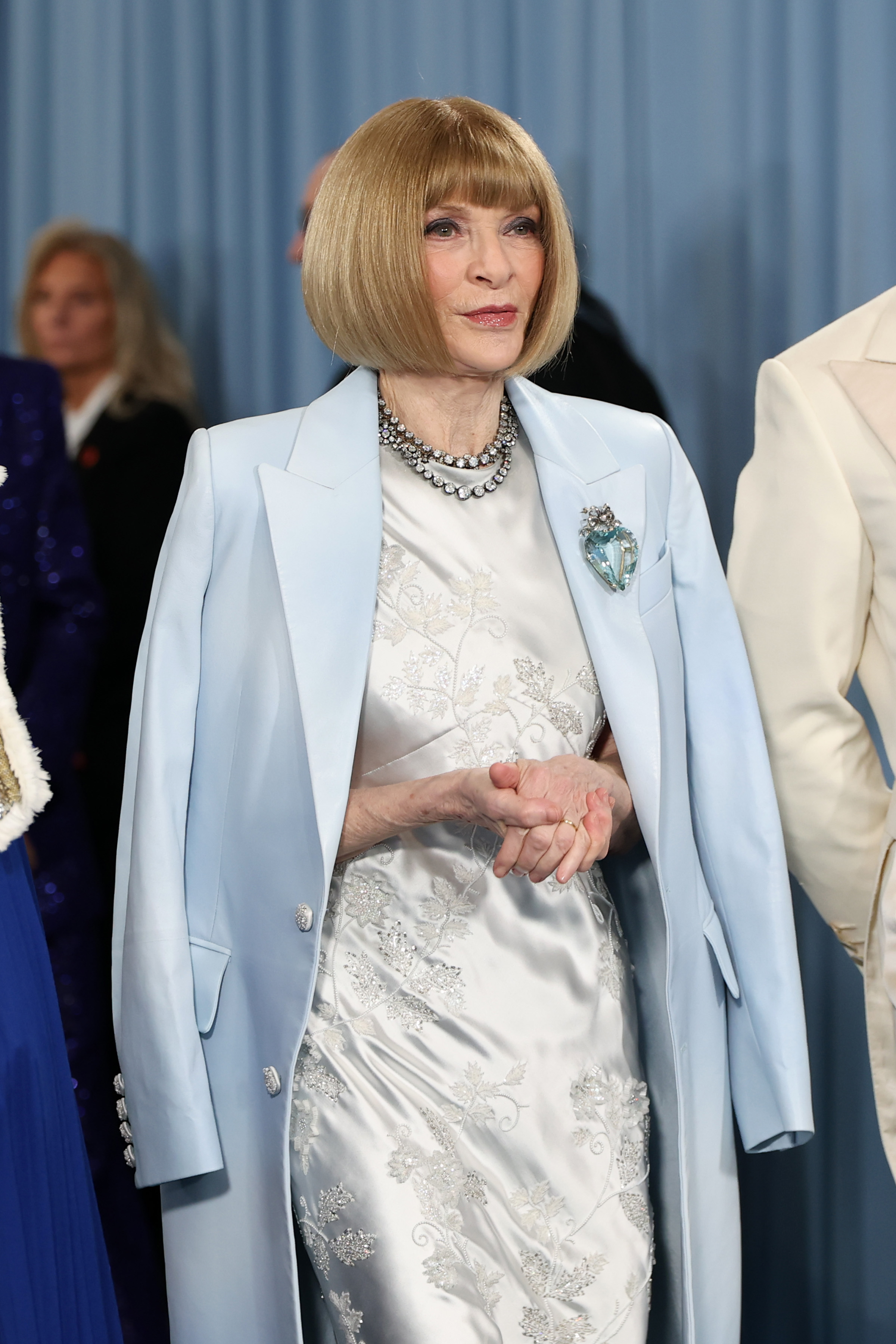Anna Wintour clasps her hands as she observes the event from inside the venue, her icy blue look adorned with a jewel brooch and statement necklace at the 2025 Met Gala honoring "Superfine: Tailoring Black Style."