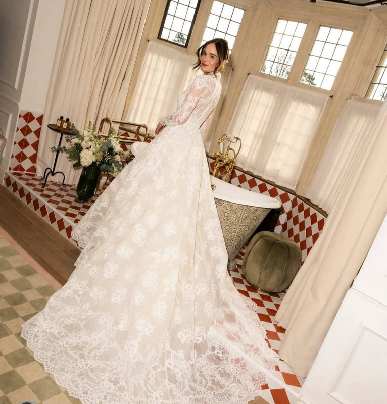 Turning towards the camera with a soft smile, Holly Ramsay stood in a tiled dressing area in her full Elie Saab gown, showing off the stunning lace train in all its detail. Her veil cascaded from a loose updo, while white florals and a vintage clawfoot tub in the background gave the scene a romantic, old-world charm. The image offered a more personal, behind-the-scenes view of the bridal preparations. | Source: Instagram/britishvogue