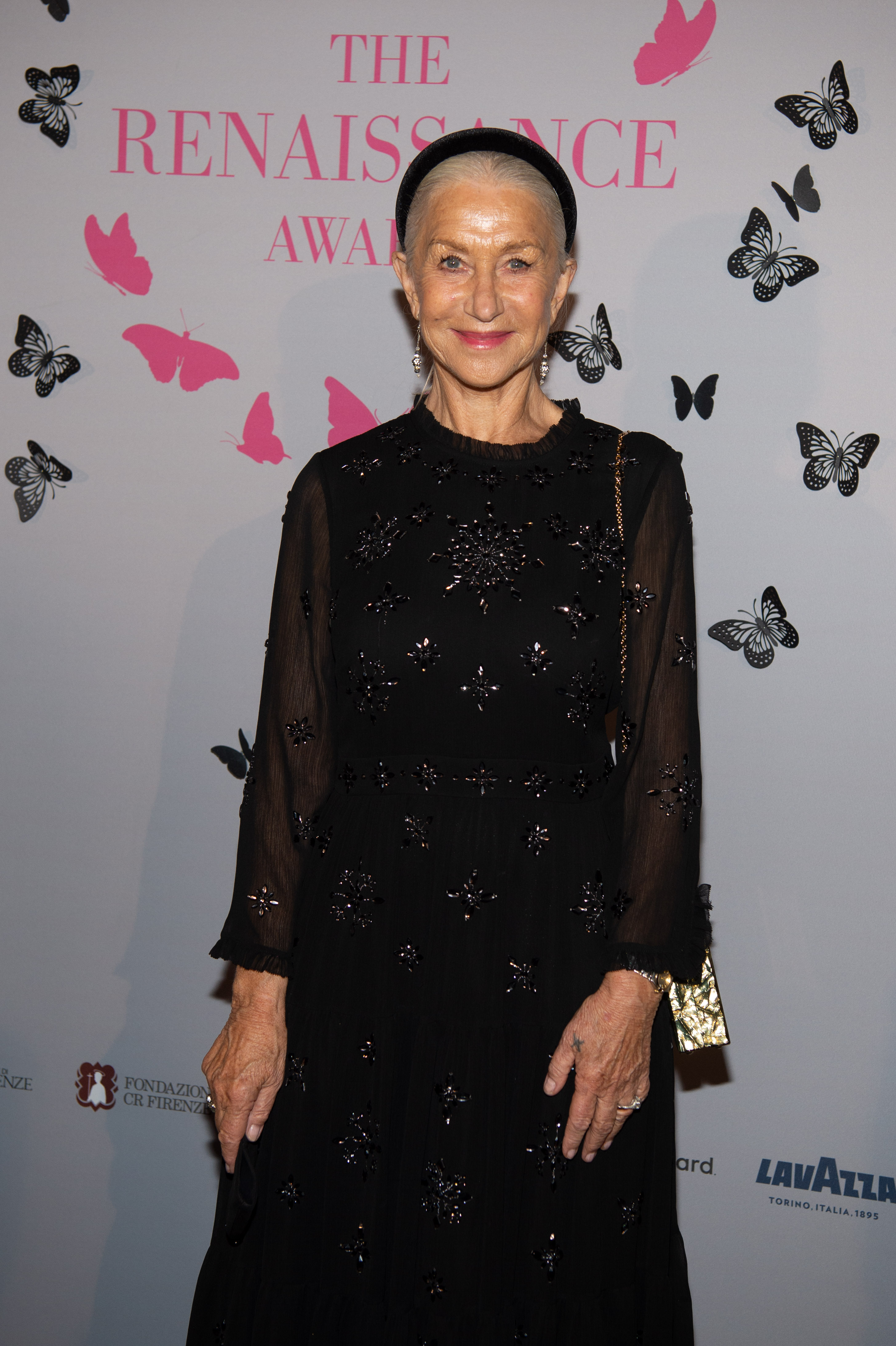 Helen Mirren attends The Renaissance Awards in a long-sleeved black gown embellished with delicate star-like beadwork, her look completed with a sleek headband, drop earrings, and a radiant smile that matches the event’s whimsical butterfly-themed backdrop.